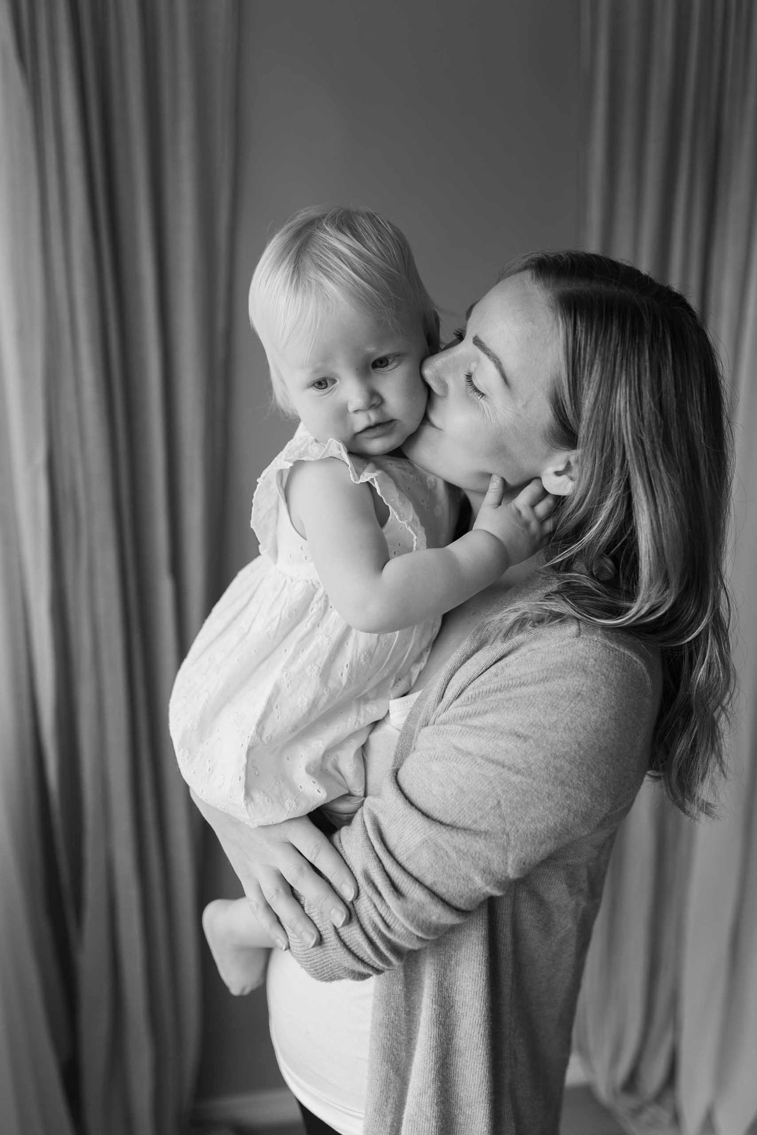 Black and white photo of a mother kissing her baby girl on the cheek while holding her in her arms.