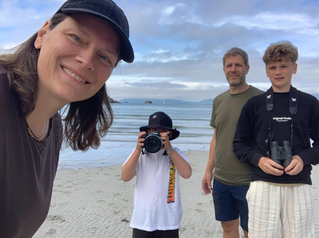 A group of photographers with cameras stand together on a beach under cloudy skies.