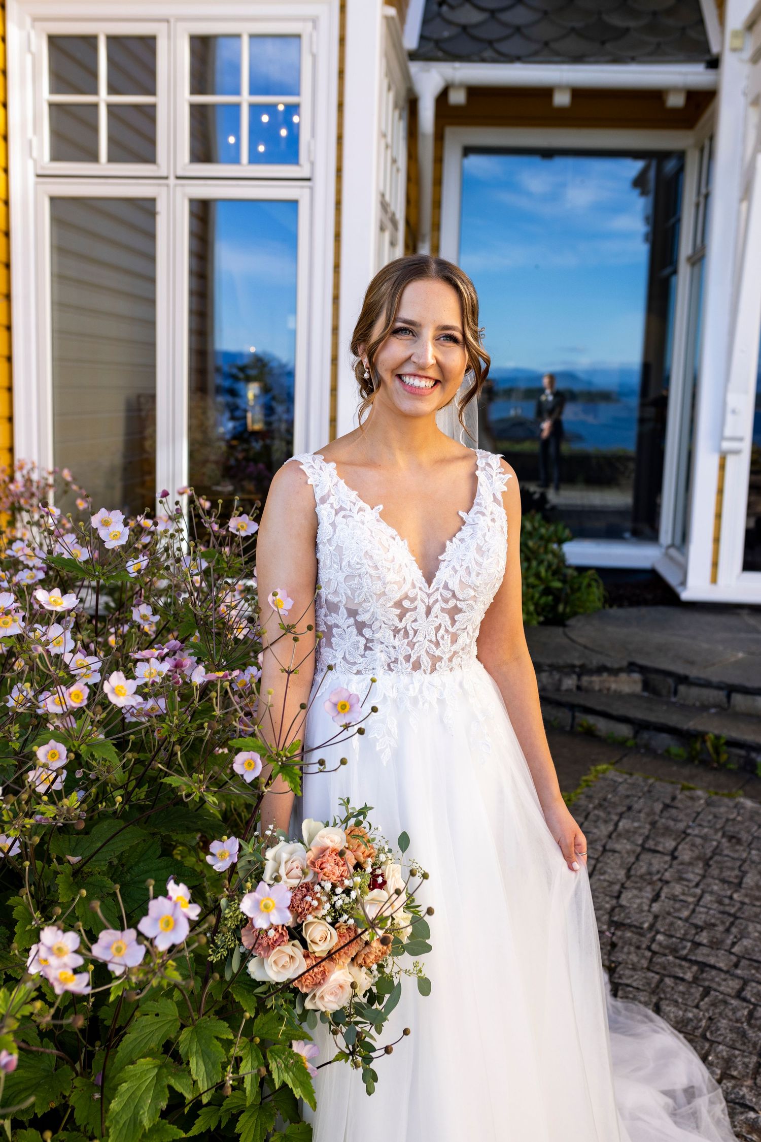Smiling bride in lace wedding dress holding bouquet of pink flowers outside a charming building on a sunny day.