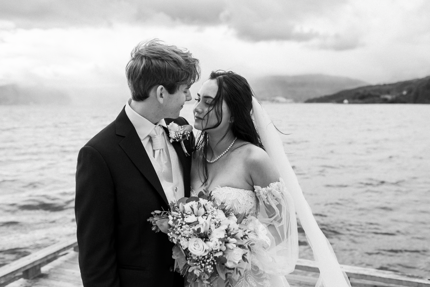 Black and white romantic wedding photo showing a couple sharing an intimate moment on a boat with water and hills behind them.
