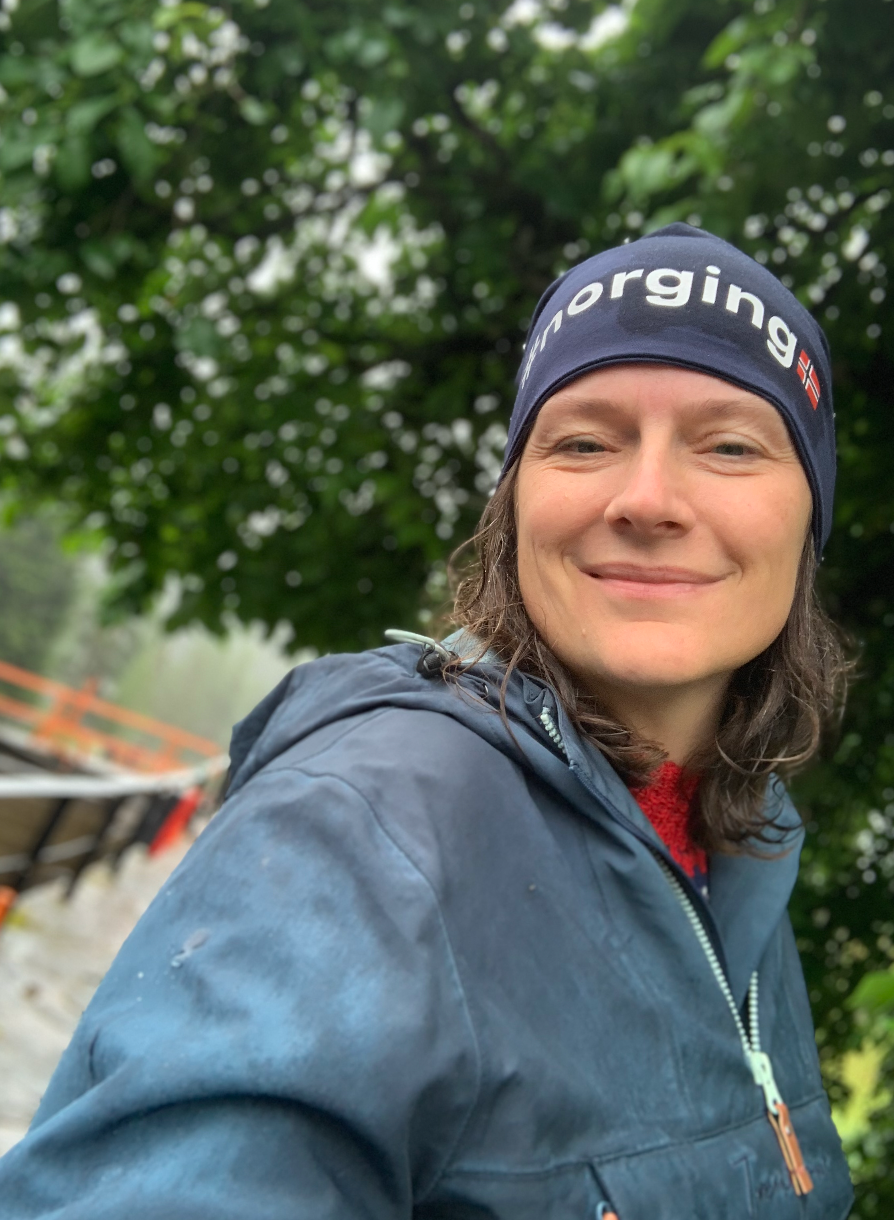 Outdoor selfie showing a person wearing a blue winter beanie and jacket against a green leafy background in nature.