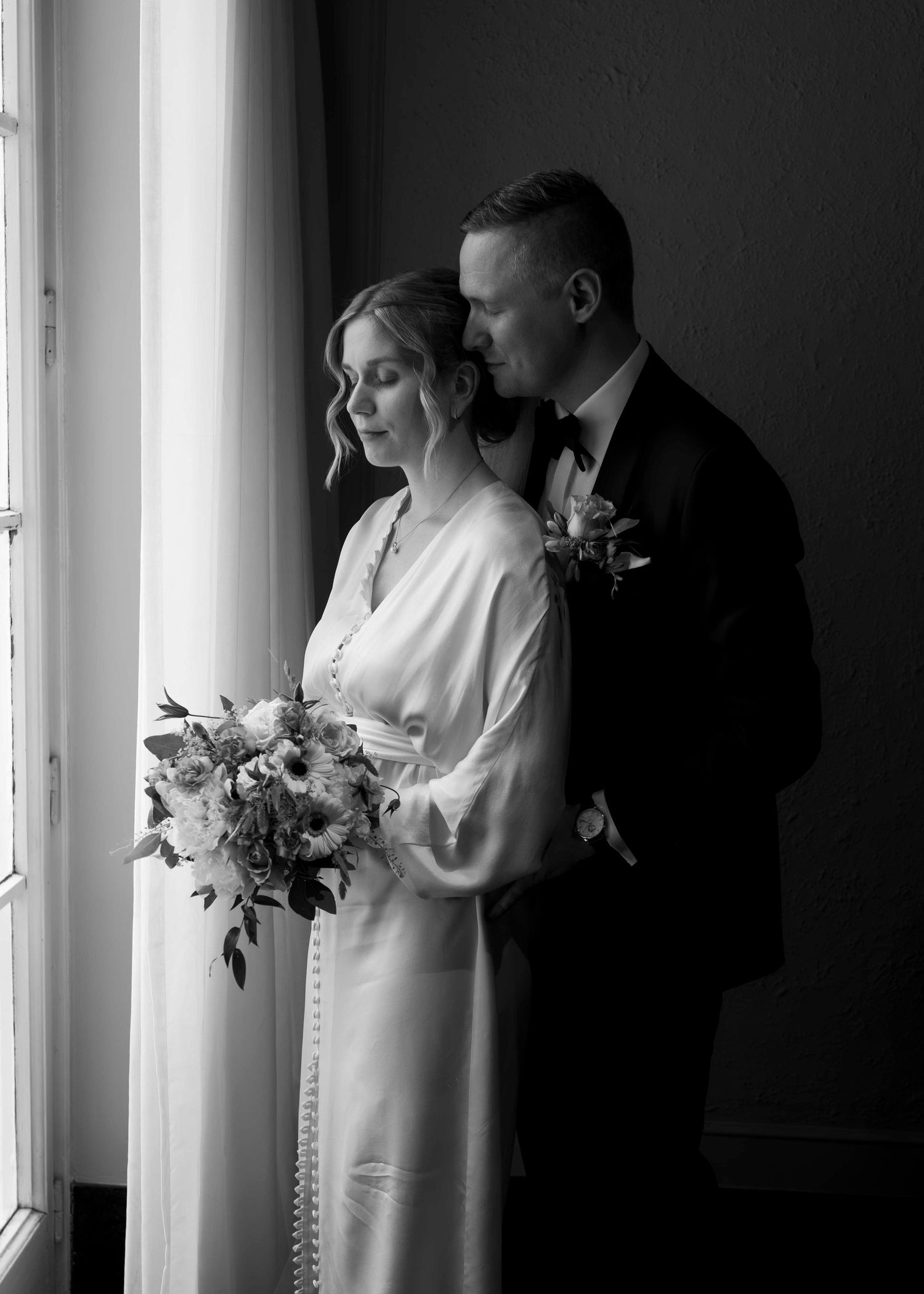 Black and white wedding photo of bride holding bouquet and groom tenderly embracing near a bright window.