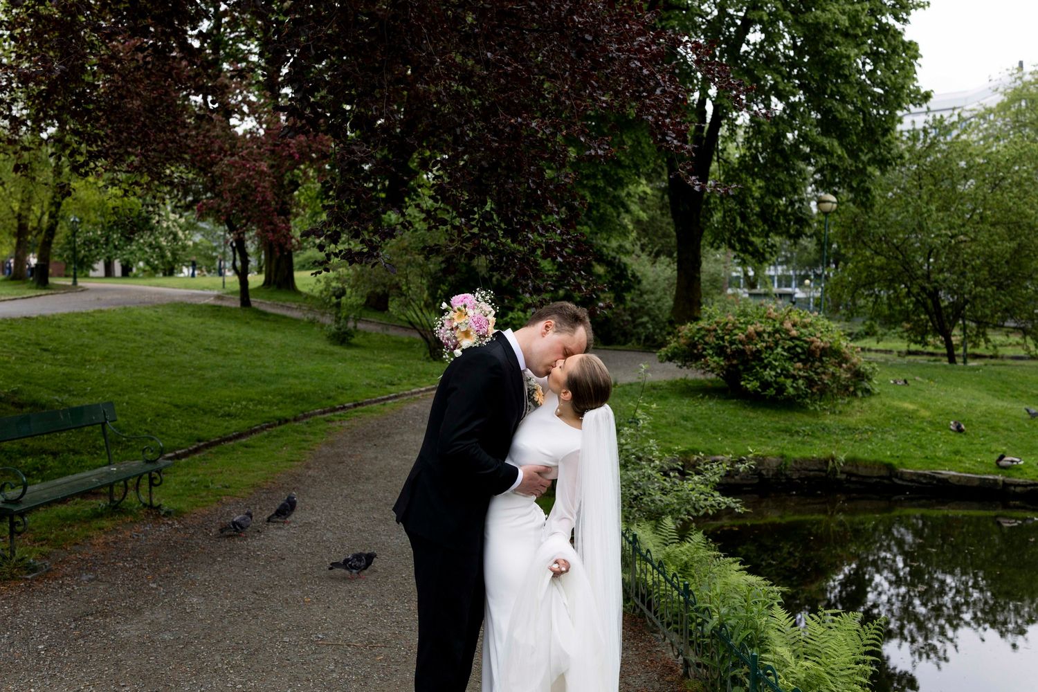 Bride and groom share a romantic kiss in a lush green park beside a pond on their wedding day.