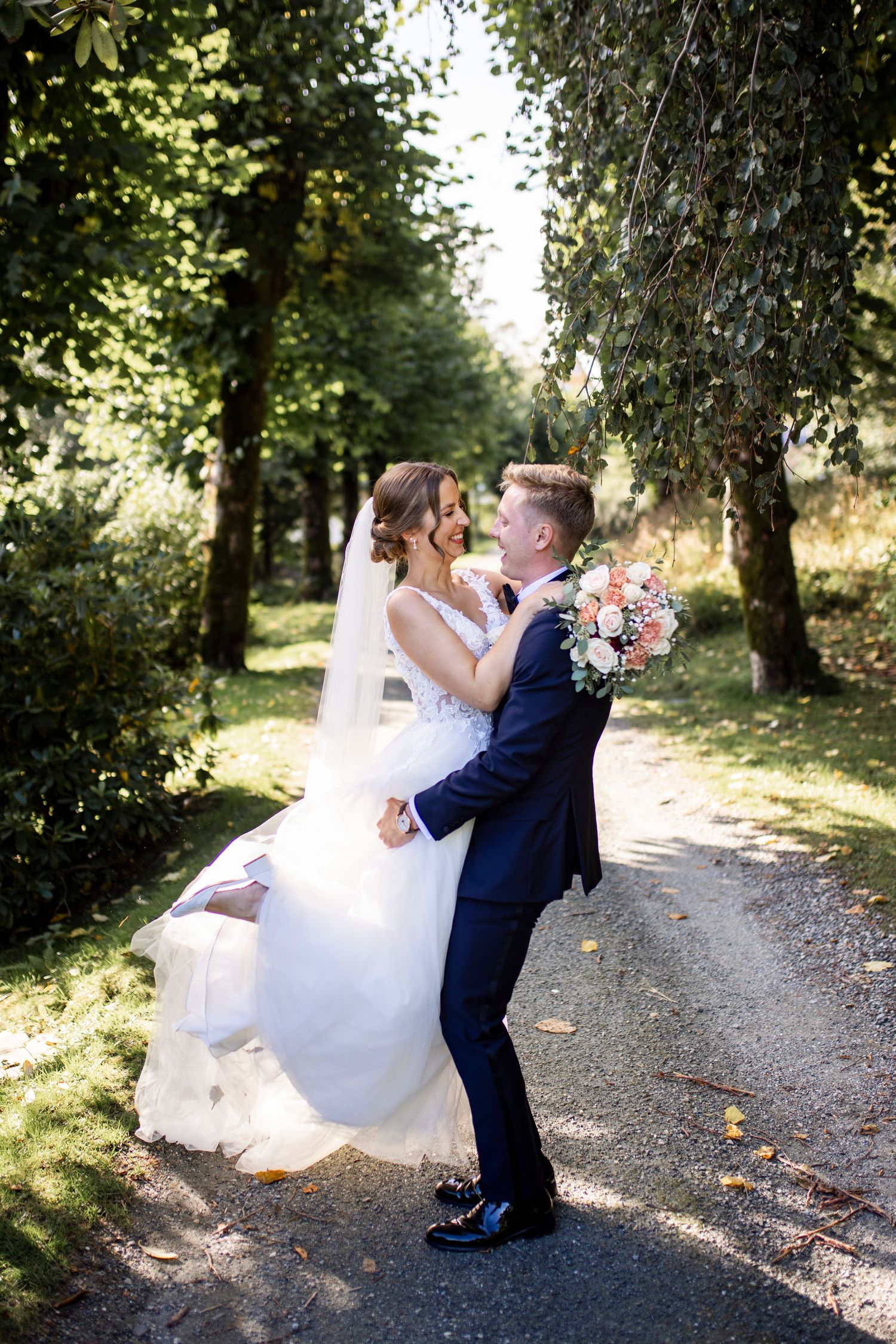 Groom in navy suit lifts bride in white wedding gown outdoors in a tree-lined park path on sunny day.