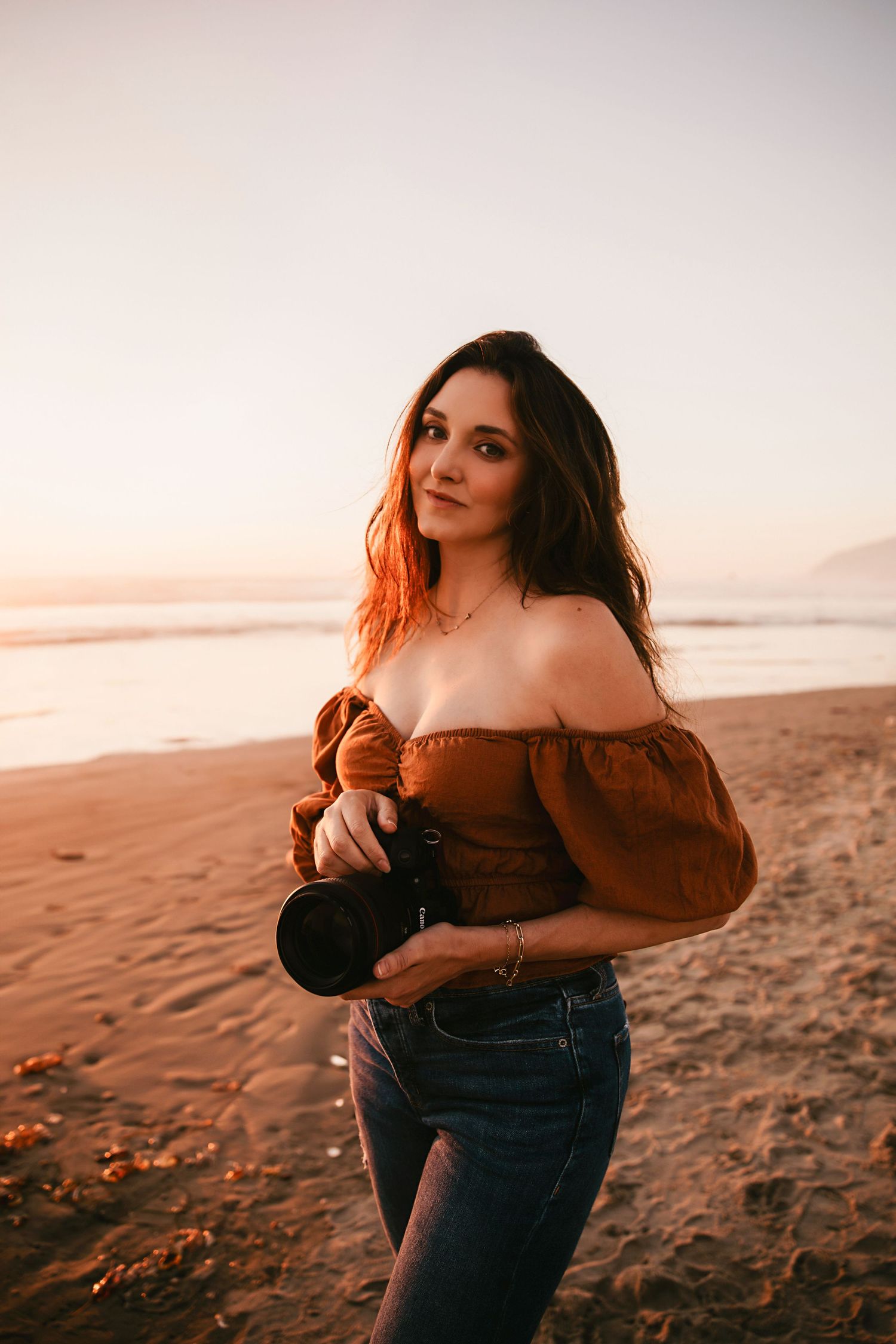Woman in brown off-shoulder top poses on beach at sunset with mountains in background.