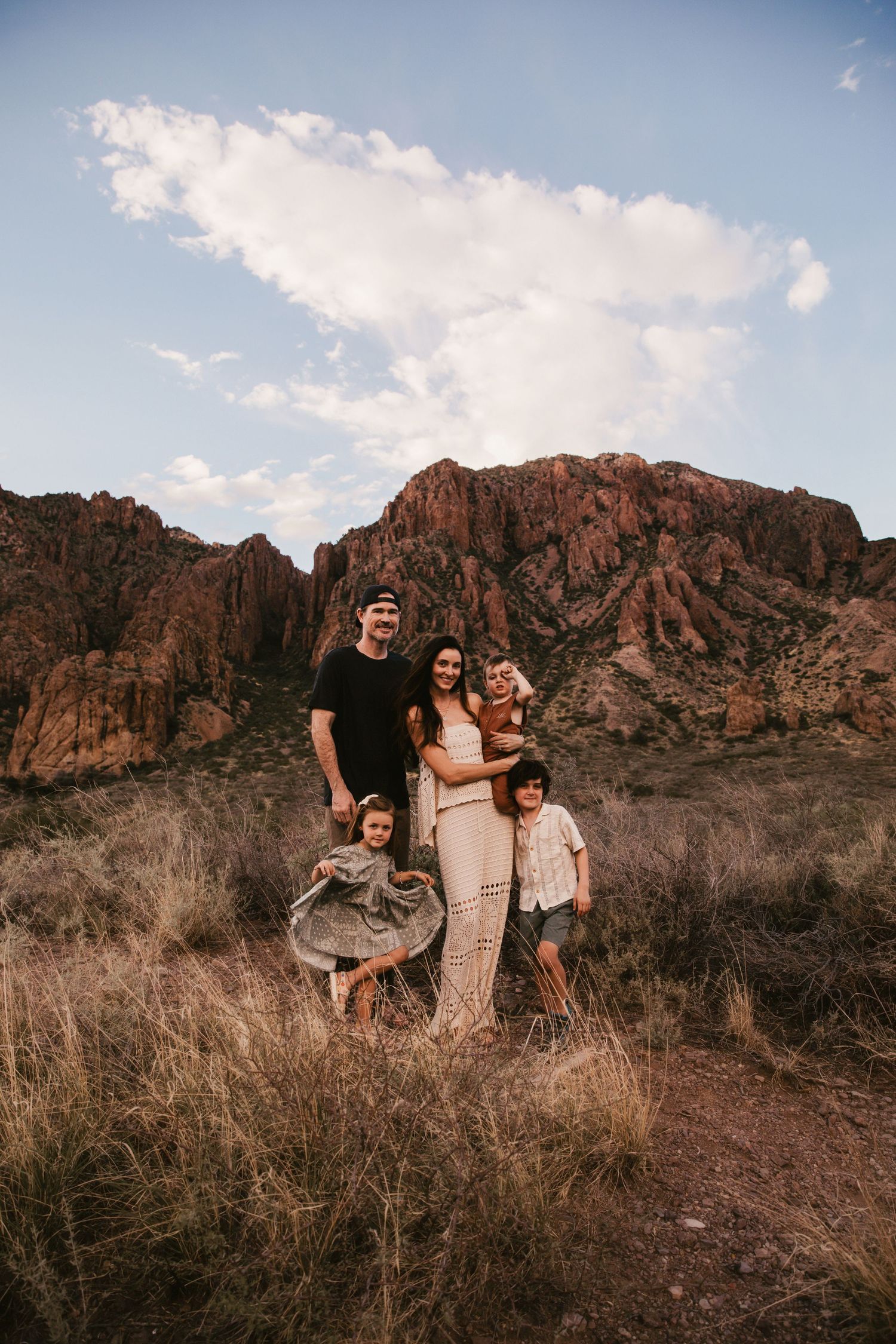 Family of five posing together in a rugged desert landscape with red rock mountains and dramatic sky backdrop.