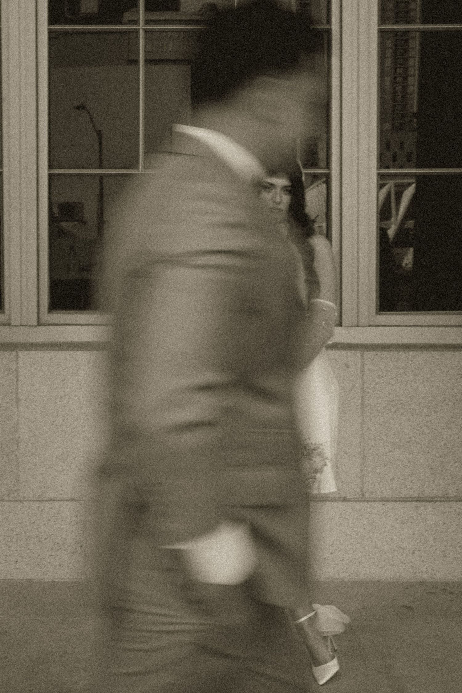 Blurred man in suit walks past a woman in white dress standing by building windows in black and white photo.