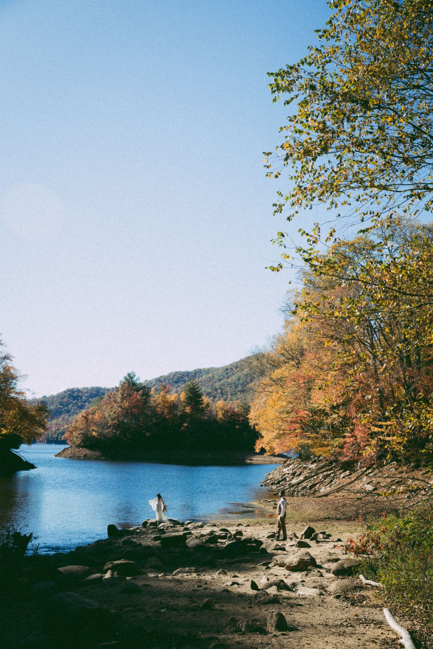 Two people stand on rocky shore of a serene autumn lake surrounded by colorful fall foliage under a clear blue sky.