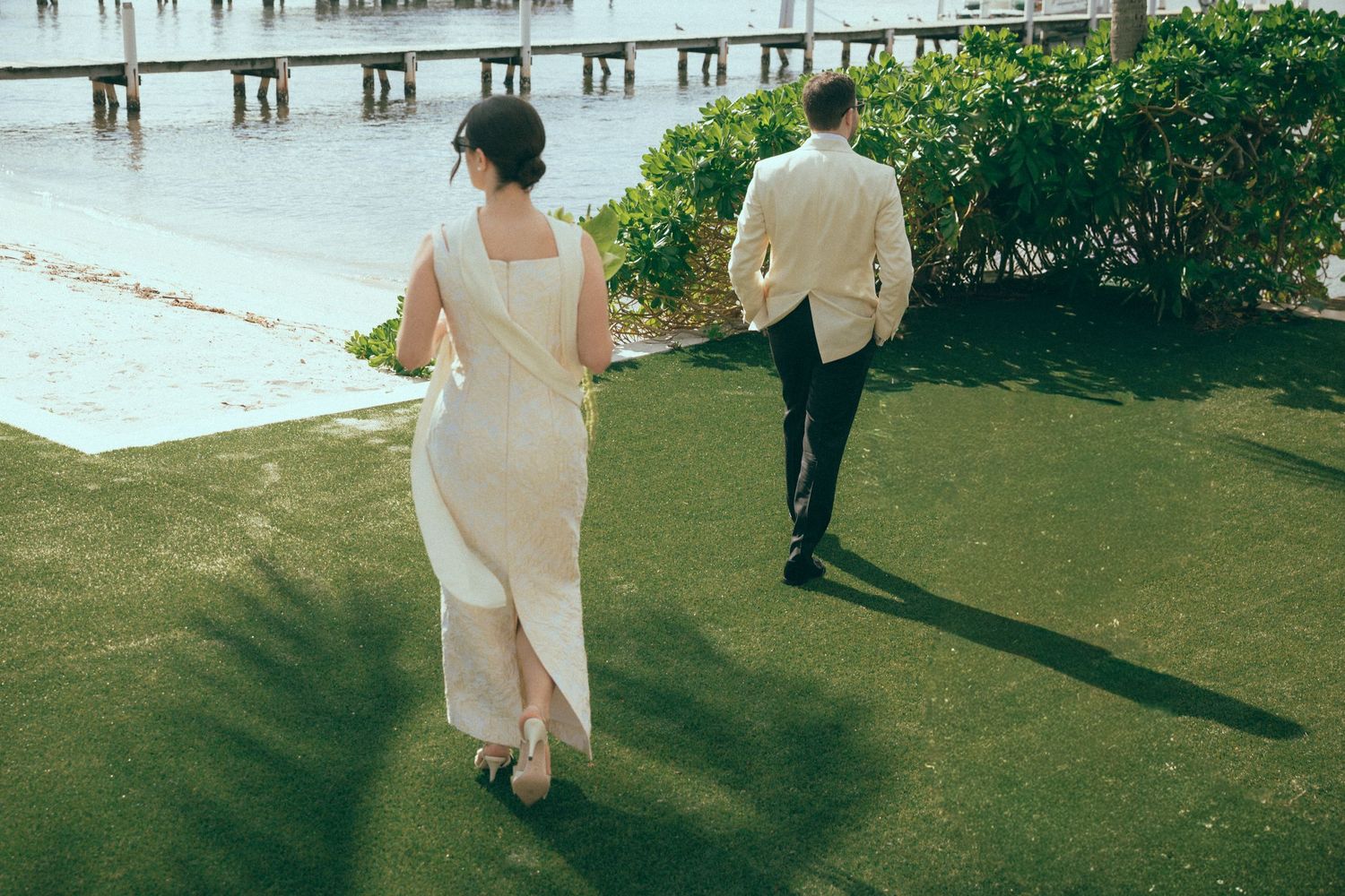 A bride and groom walk hand in hand on green grass by a waterfront dock on their wedding day.