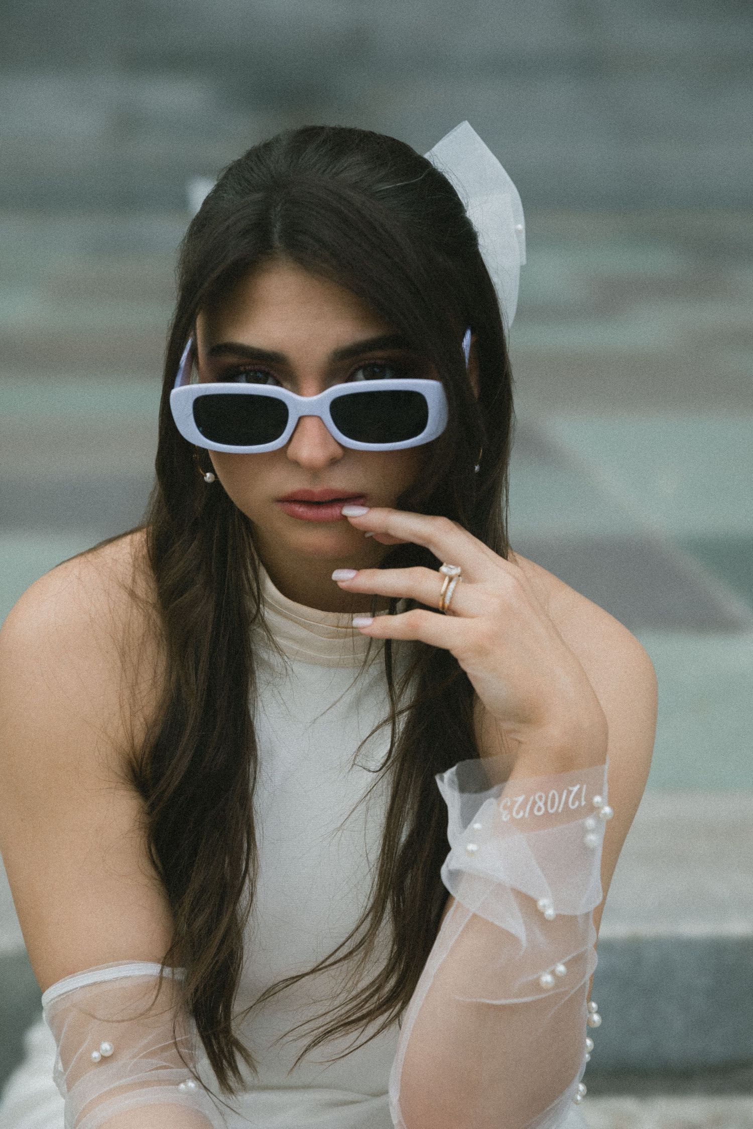 Young woman in white sunglasses, sheer gloves with pearls, and bow hair accessory posing on steps.