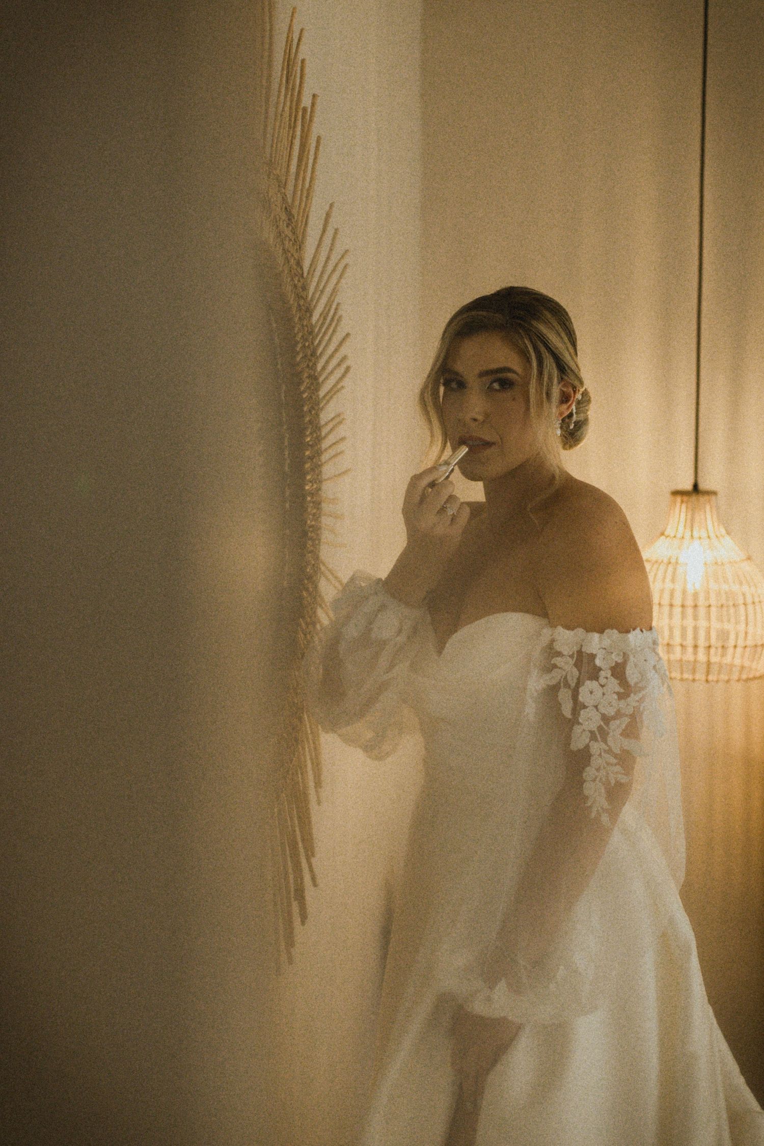 Bride in off-shoulder white gown with floral lace sleeves applying lipstick in warm ambient lighting.