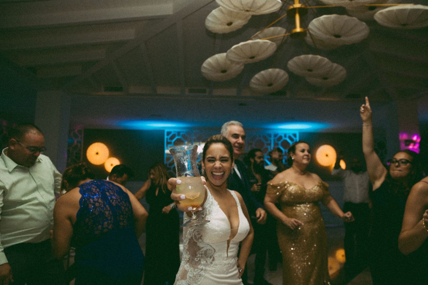 Bride in white dress holding large drink glass dancing joyfully at wedding reception with guests celebrating on dance floor.