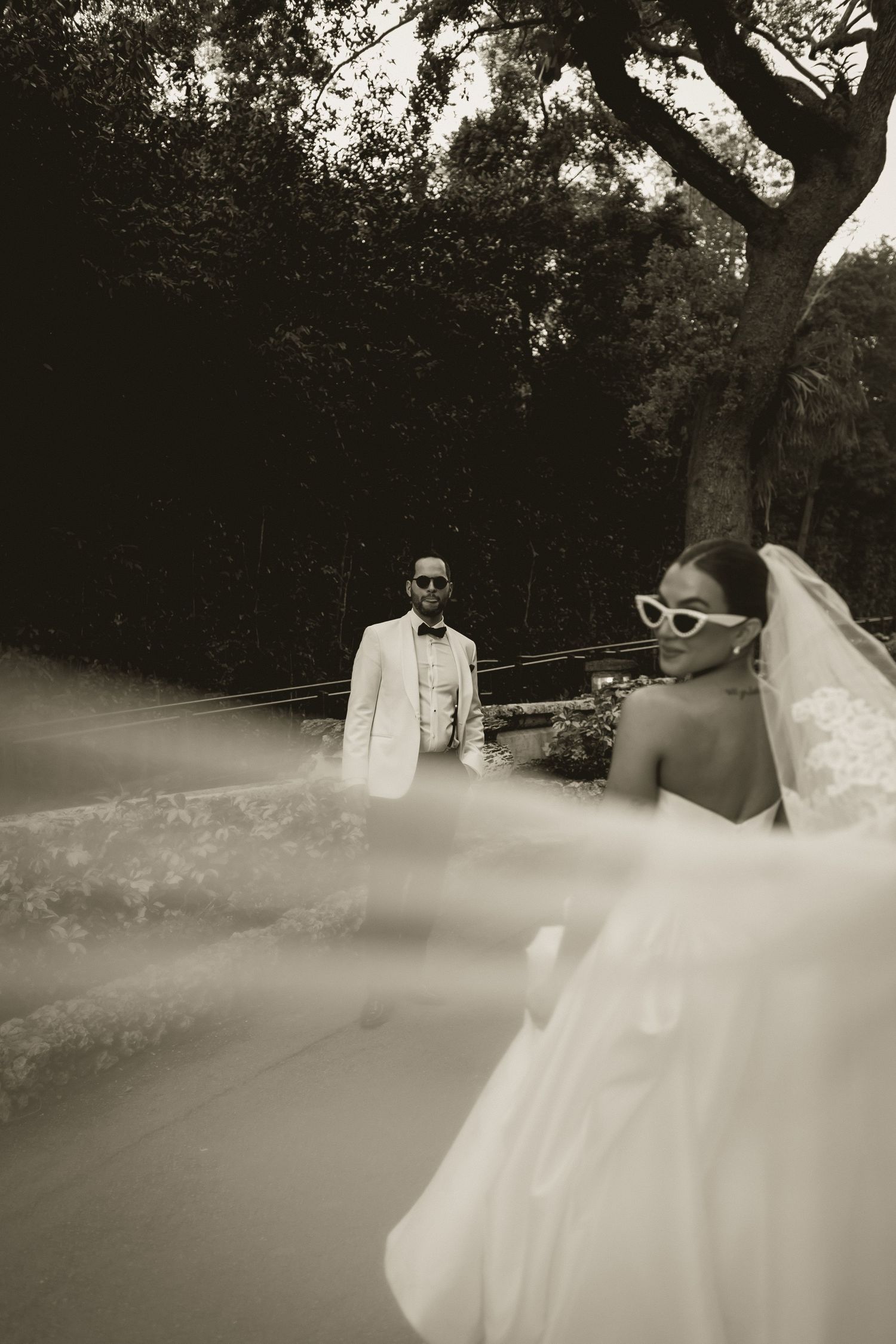 Black and white wedding photo of bride in flowing veil and sunglasses with groom in white suit outdoors.