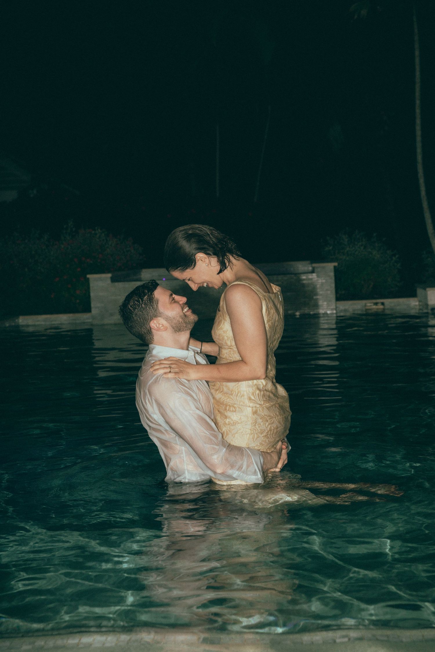 Romantic couple embracing in a pool at night, wearing formal attire, sharing an intimate moment.