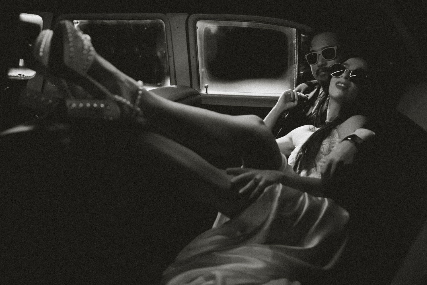 Black and white photo of a couple relaxing inside a vintage car, woman resting legs on dashboard.