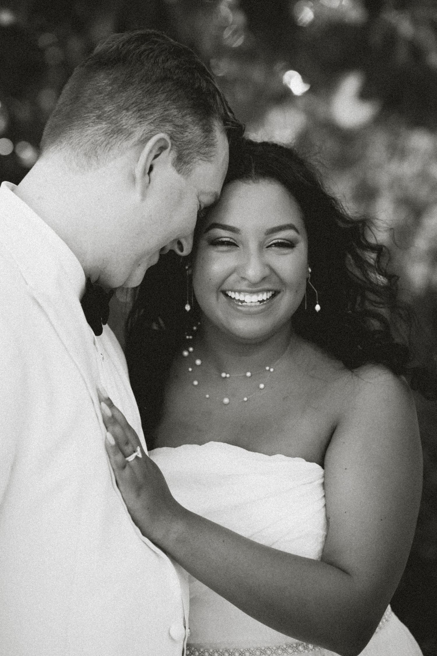 Black and white photo of a smiling bride in strapless gown leaning close to groom in white shirt outdoors.
