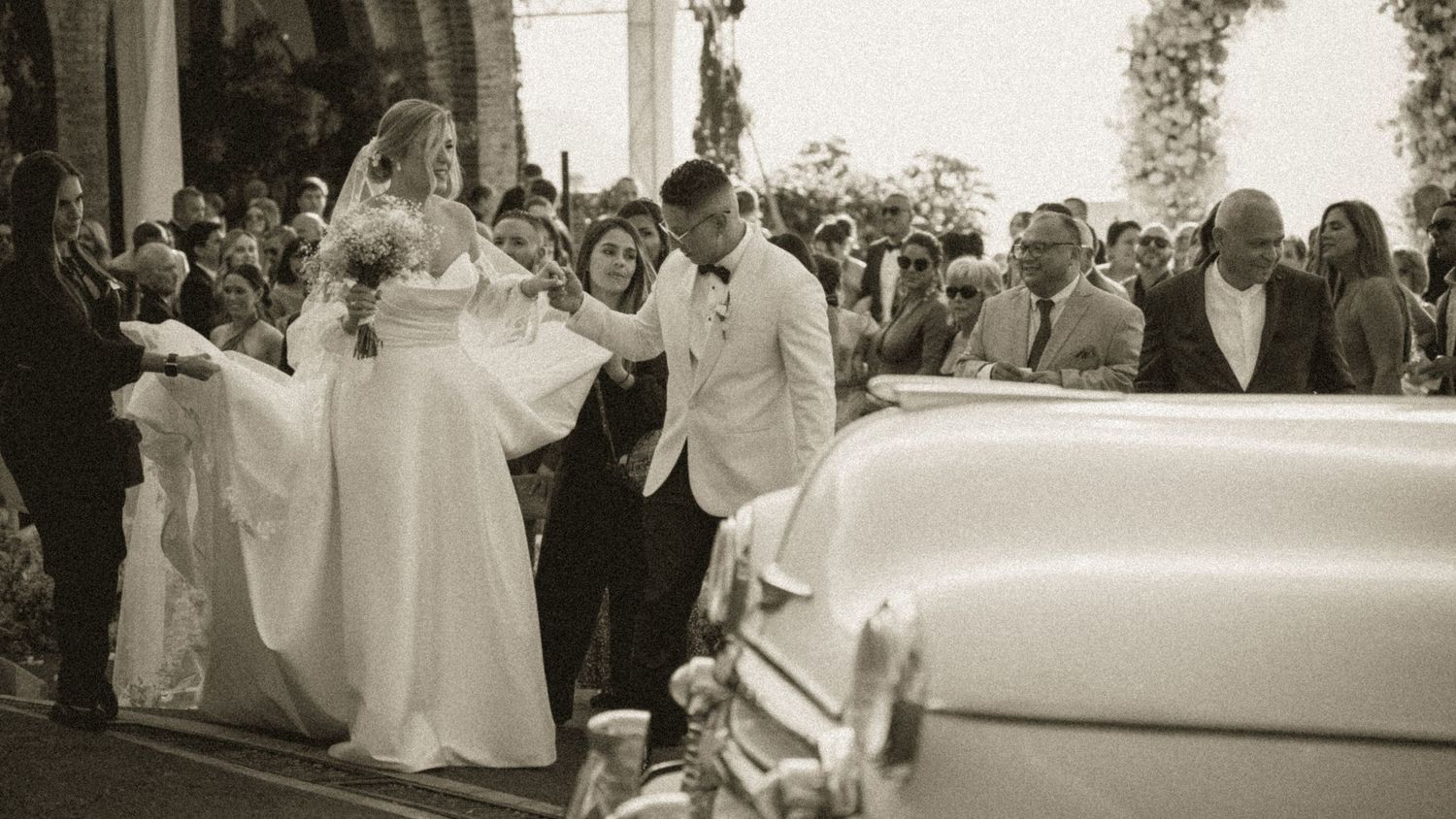 A bride in a white gown and groom in a white suit exit a church ceremony beside a vintage car surrounded by guests.
