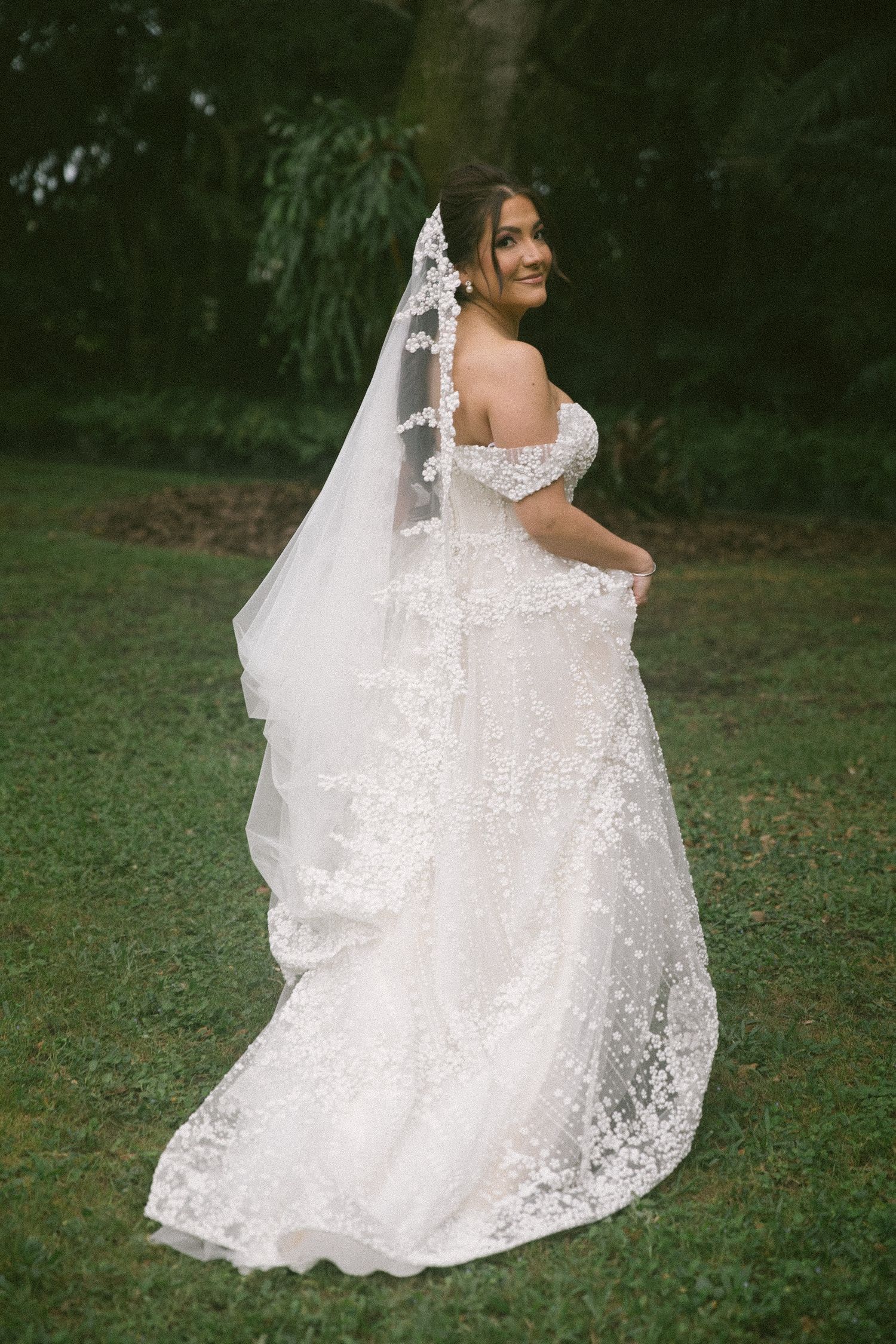 Bride in off-shoulder lace wedding gown with cathedral veil smiling over shoulder in lush garden setting.