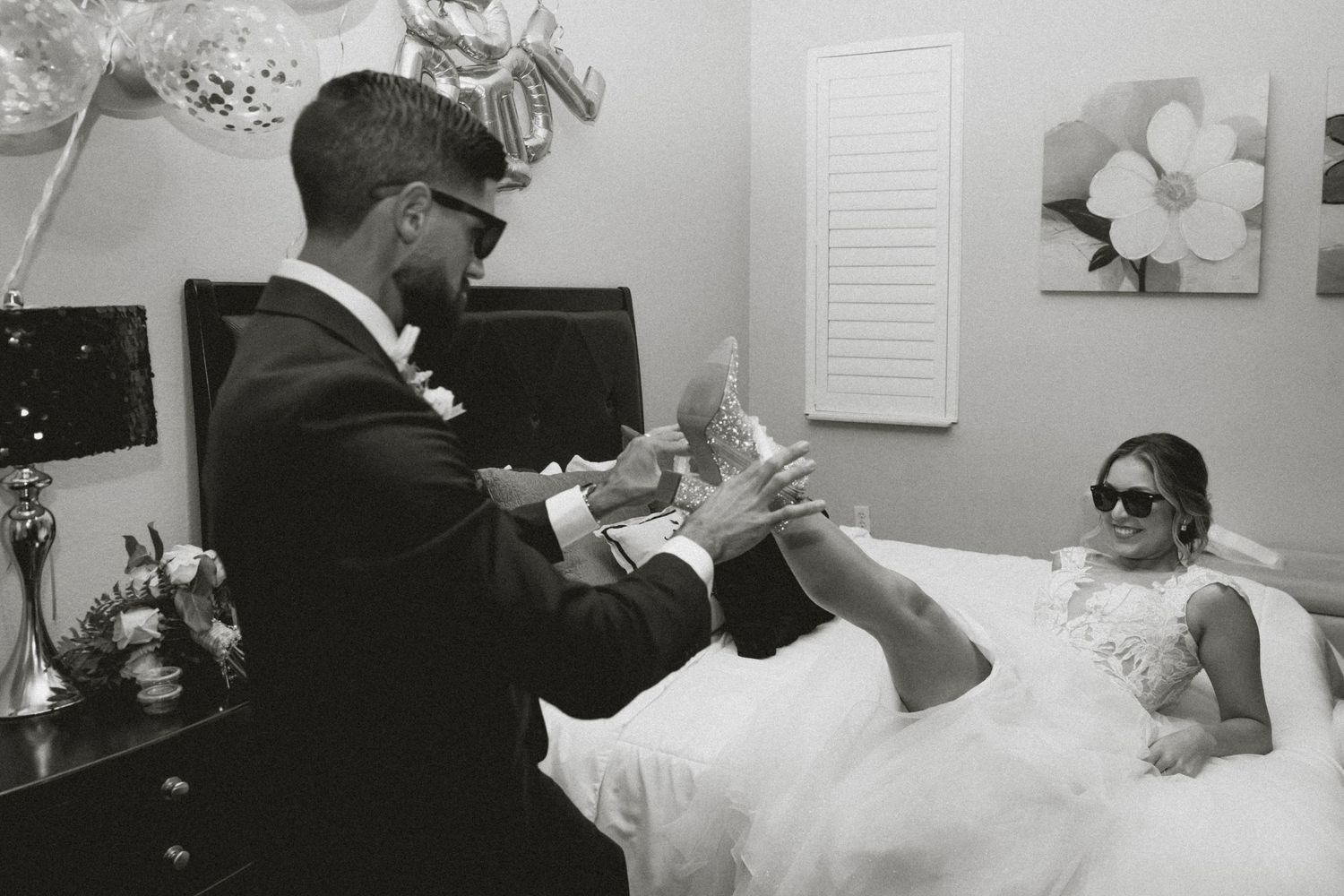 Black and white wedding photo of groom adjusting brides shoe while she reclines, both wearing sunglasses indoors.
