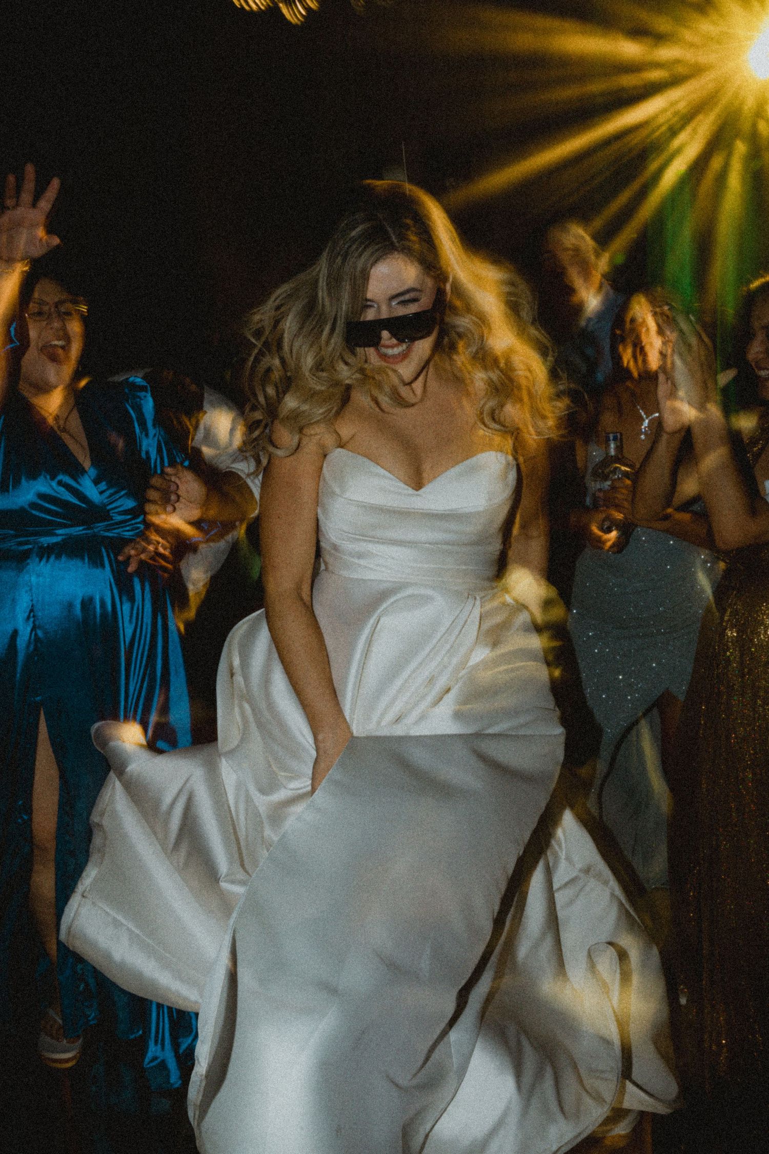 Bride in white strapless gown and sunglasses dancing energetically at wedding reception with colorful stage lights.