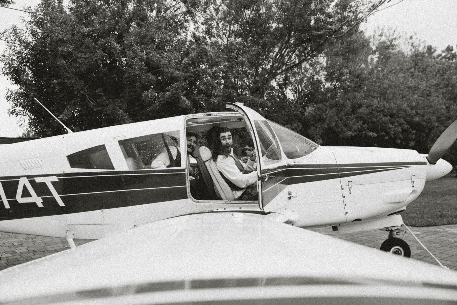 Vintage black and white photo of a person sitting in a small private plane cockpit, with trees visible in the background.