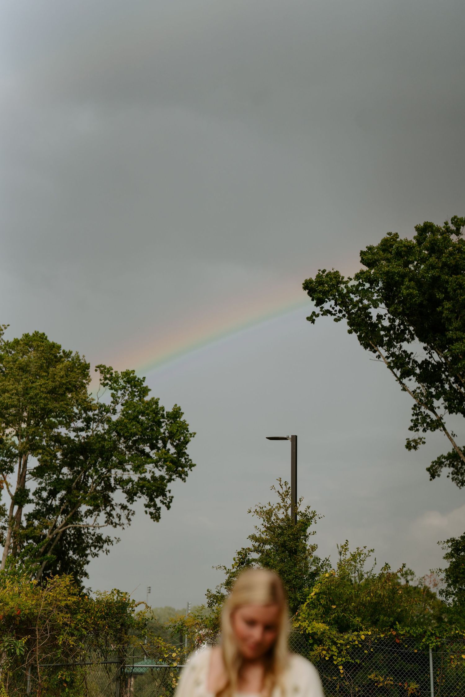 Blonde woman stands in foreground while vibrant rainbow arcs across stormy gray sky above green trees.
