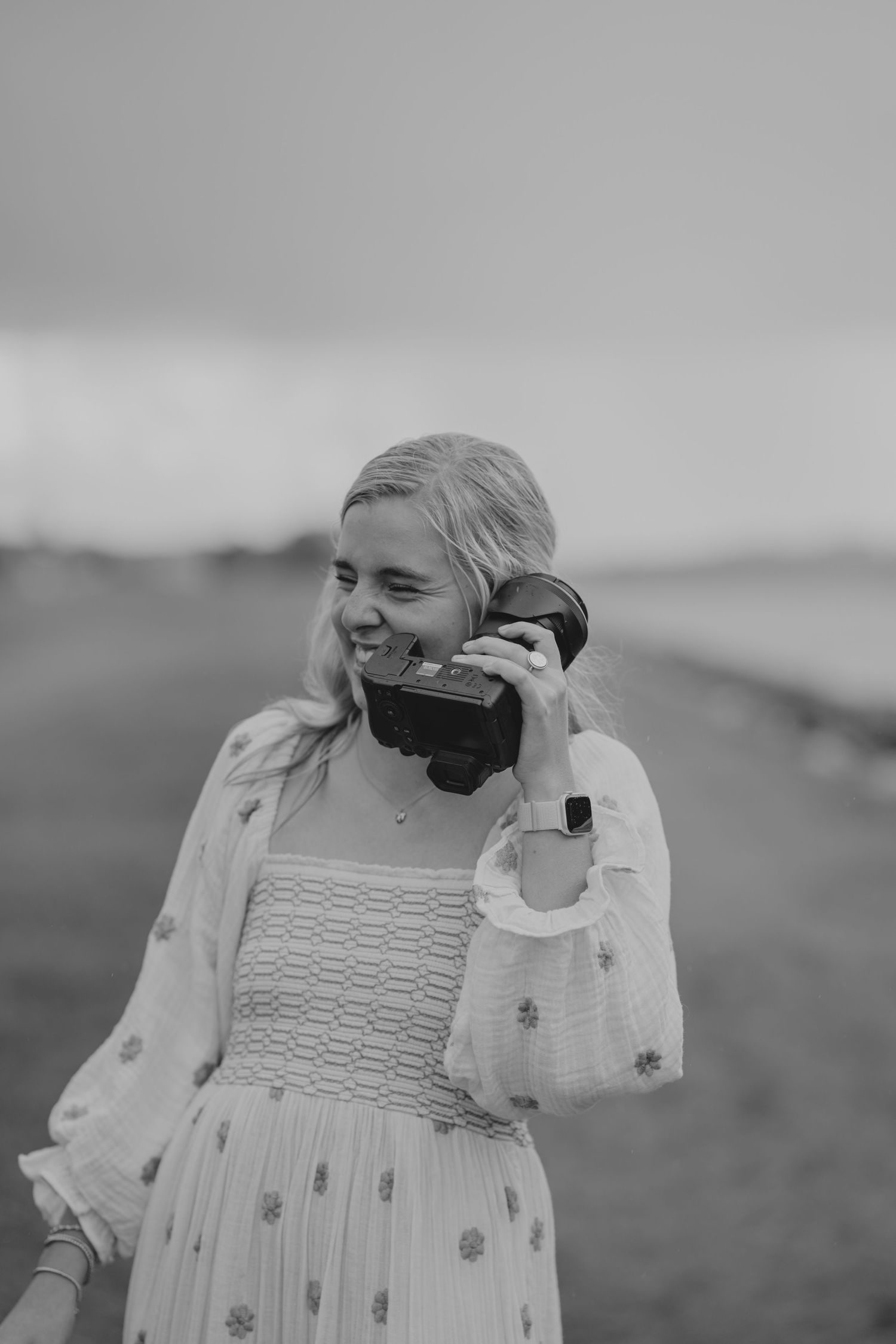 Smiling blonde woman in white floral dress holds vintage film camera near her face in black and white photo.