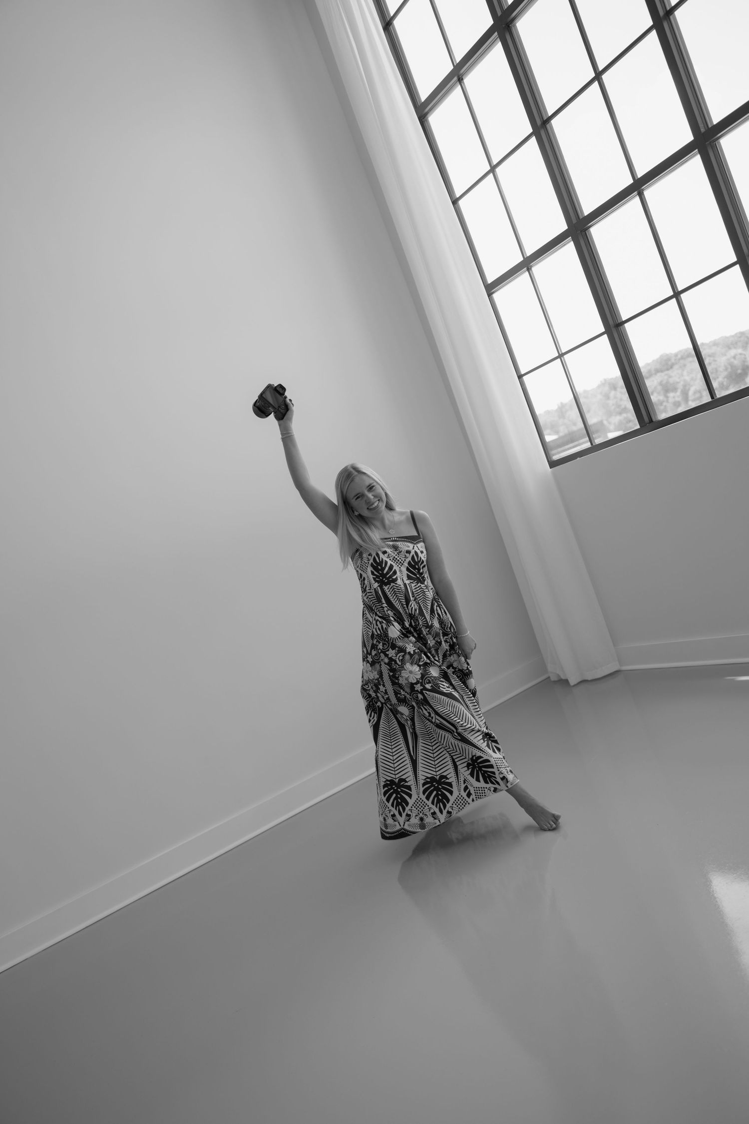Woman in floral maxi dress holding camera aloft in bright minimalist studio with large windows, black and white photo.