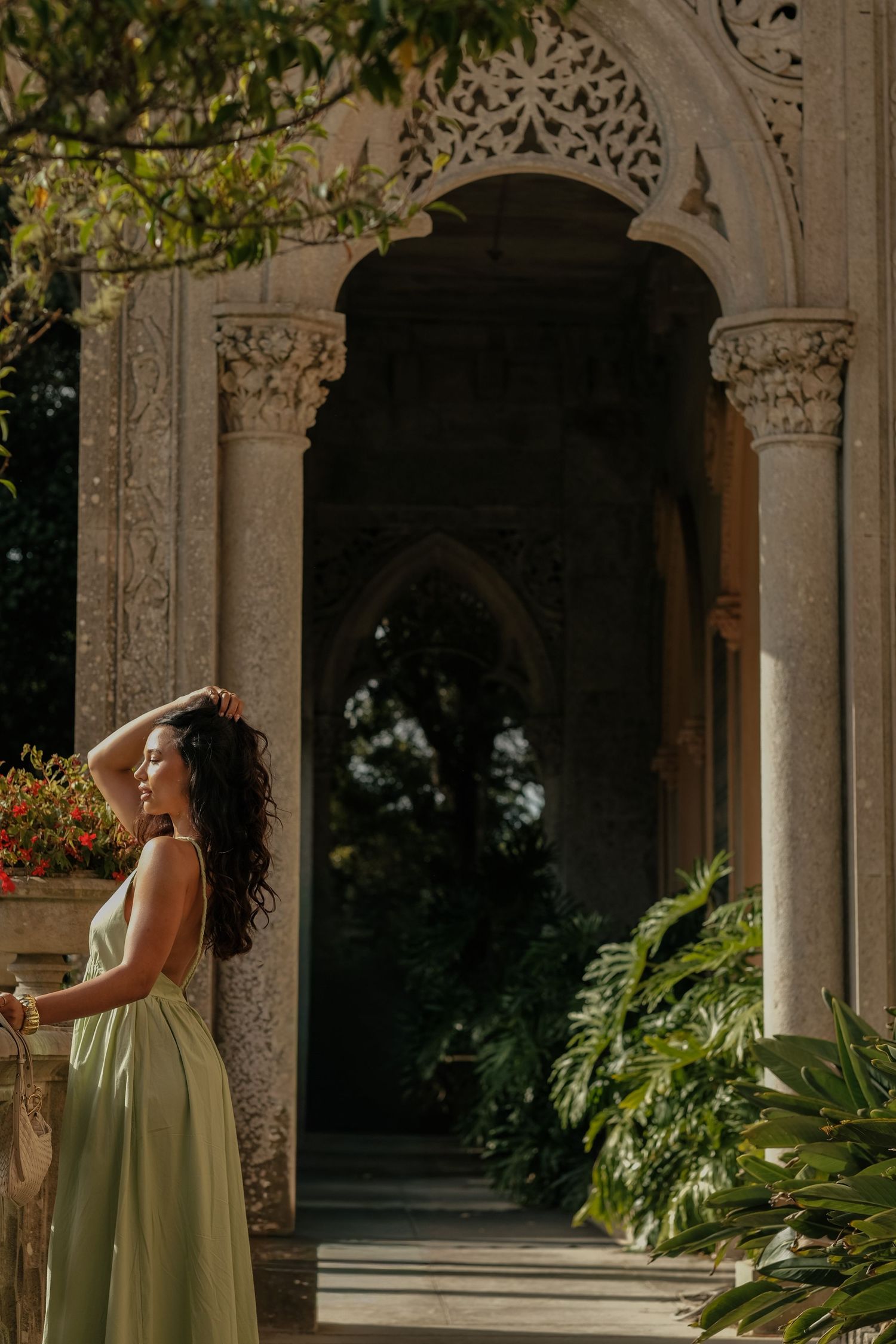 Woman in sage green dress poses elegantly among ornate stone archways and lush greenery in a classical garden setting.
