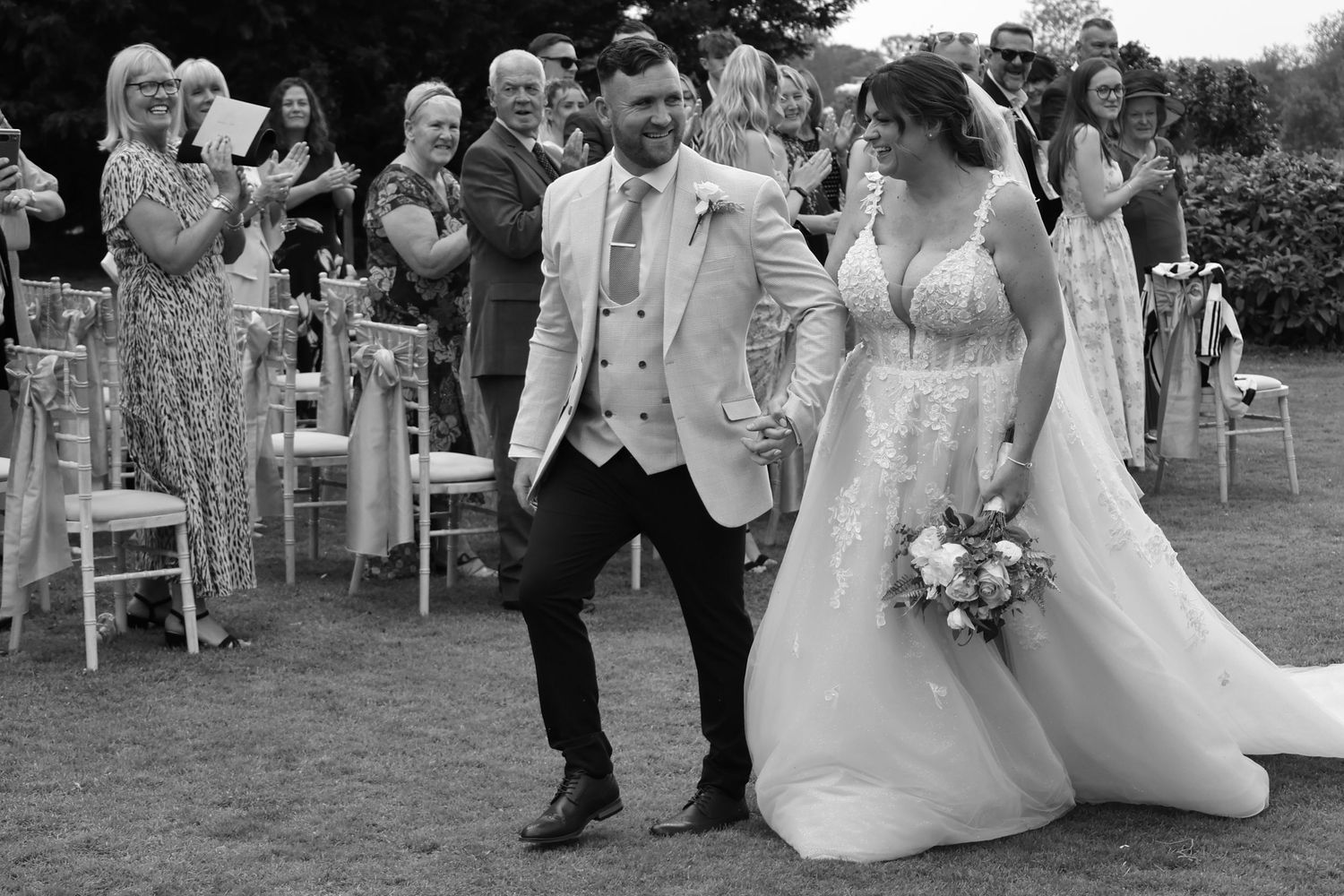 Black and white photo of a joyful newlywed couple walking hand in hand at an outdoor wedding ceremony.