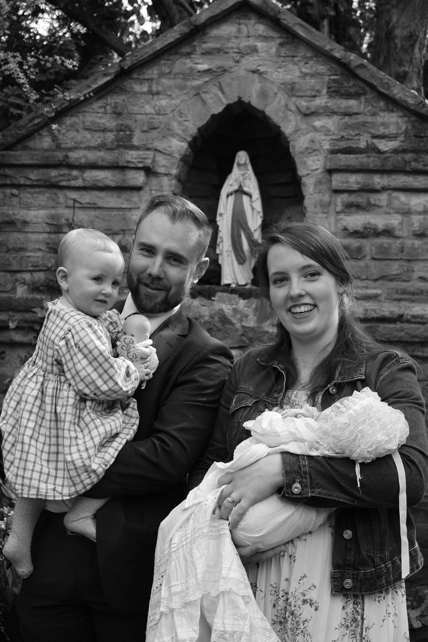 A family poses in front of a stone grotto with a statue of the Virgin Mary for what appears to be a christening or baptism.