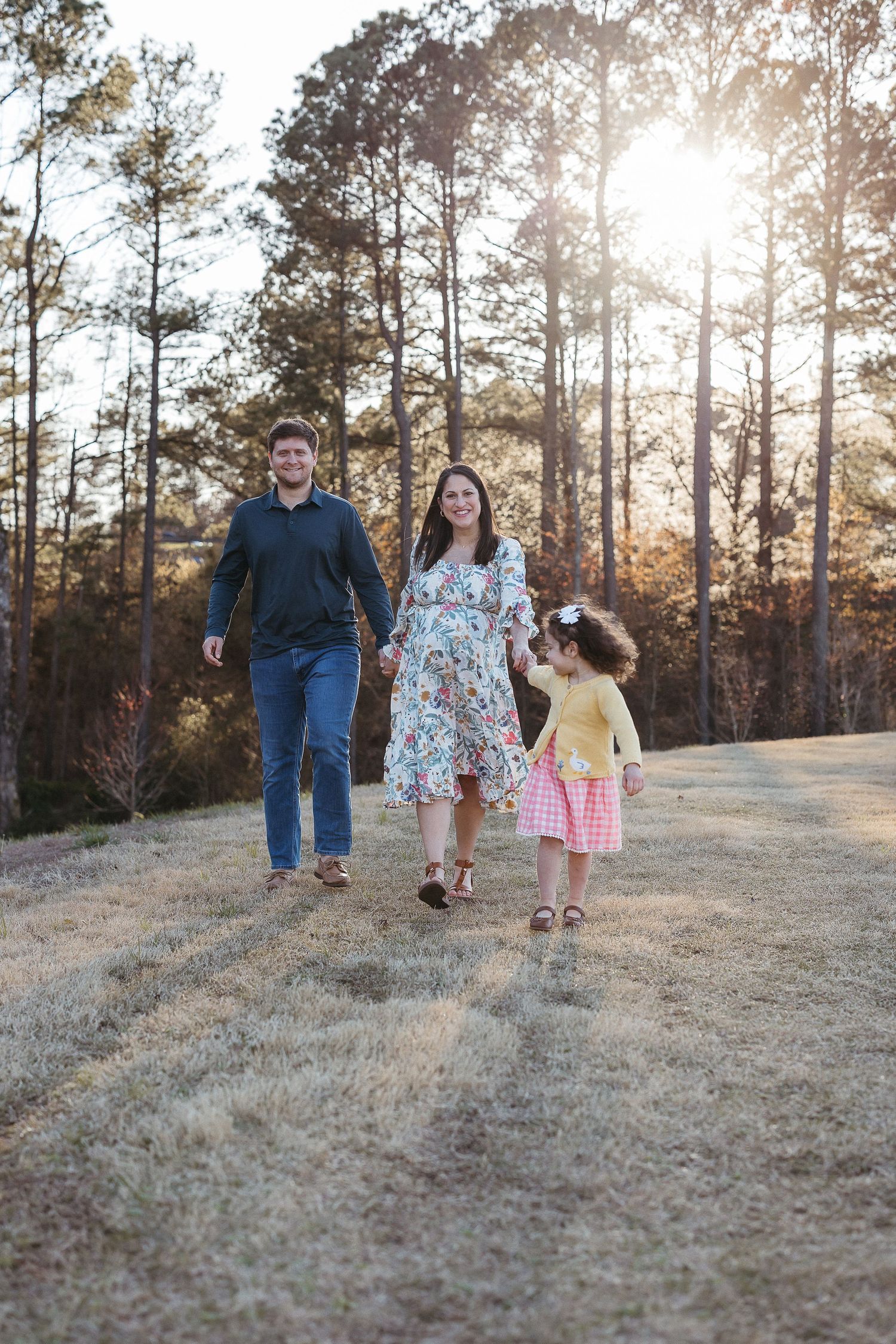 Three people walking on a dirt path through pine woods at golden hour with sunlight filtering through trees.