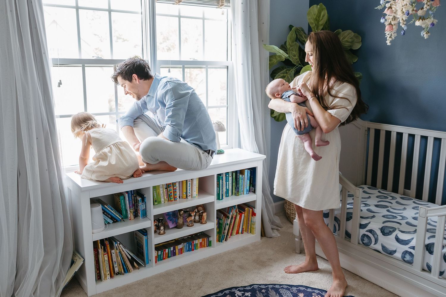 Parents with baby in cozy nursery with built-in window seat bookshelf and navy blue accent wall.