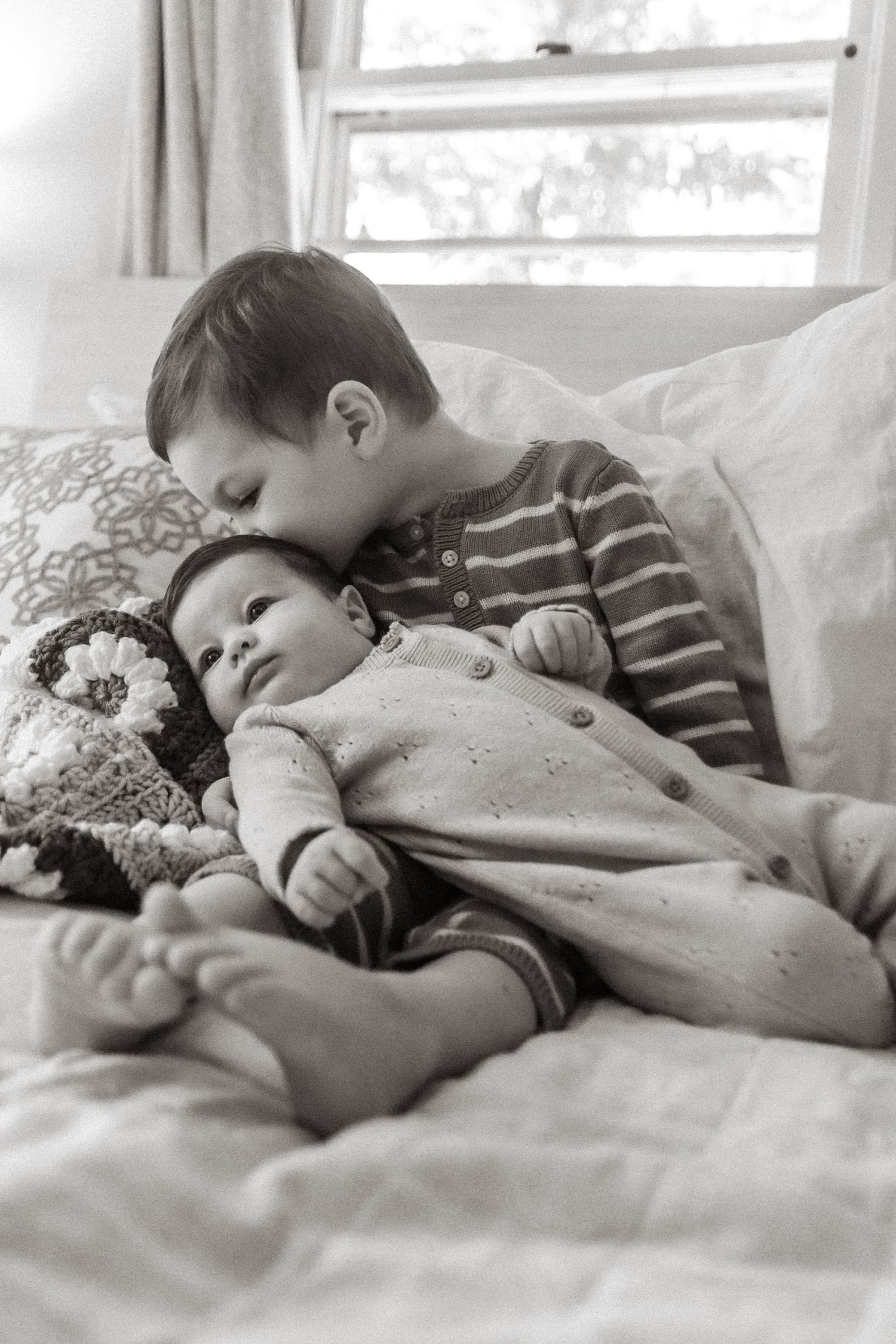 A tender black and white photograph showing two siblings resting together on a bed with a stuffed animal.
