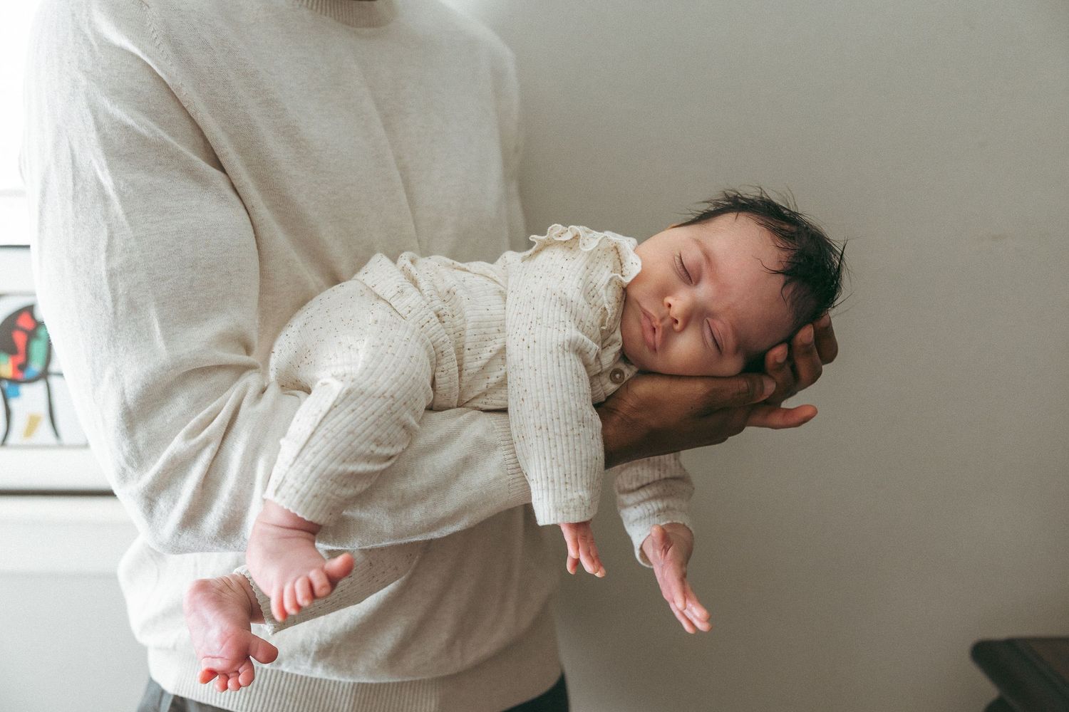 A sleeping baby wearing a white onesie is cradled in someone's arms against a light gray background.