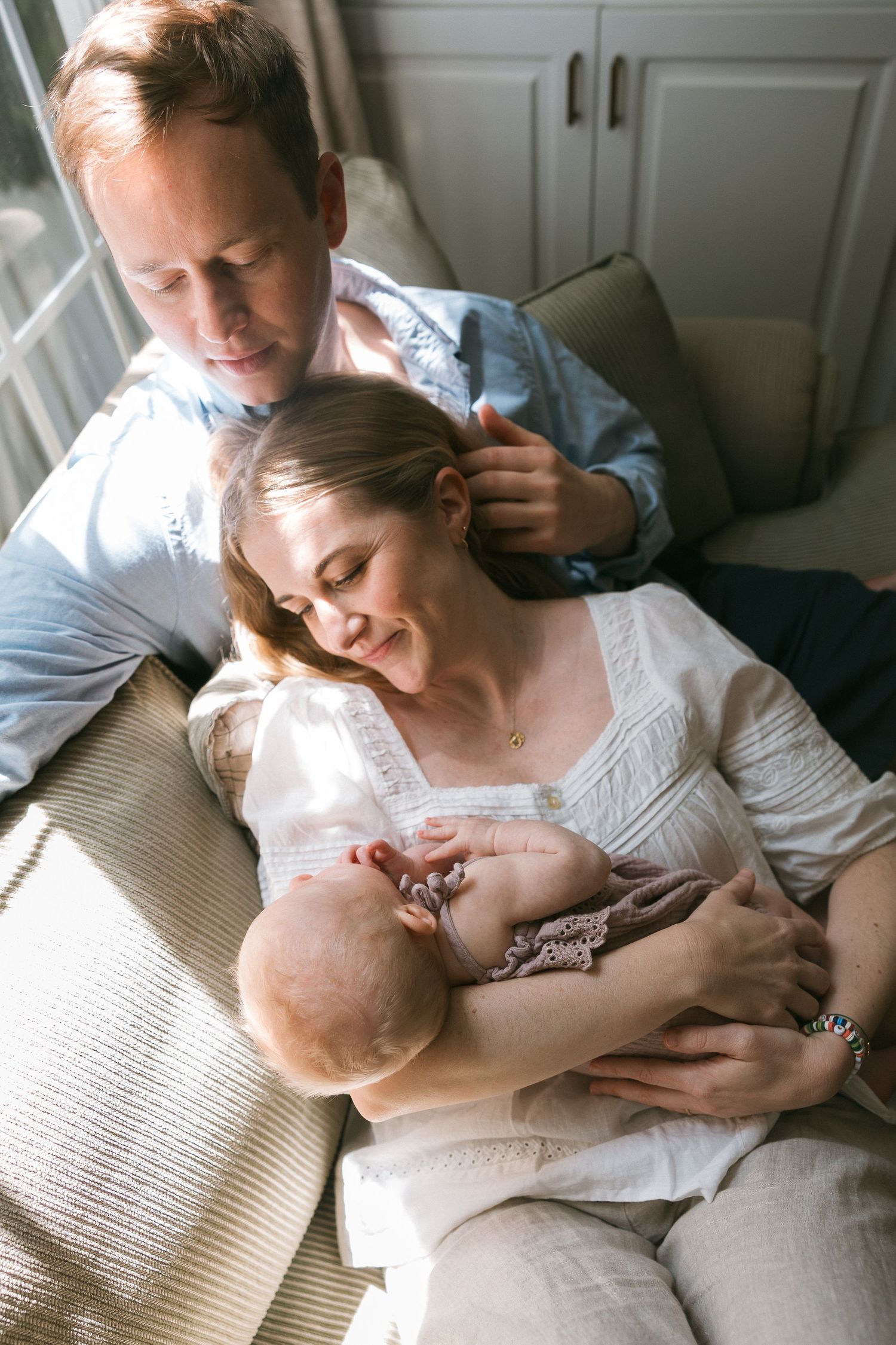 A couple sits together on a couch, smiling as they cradle their newborn baby in a warmly lit room.
