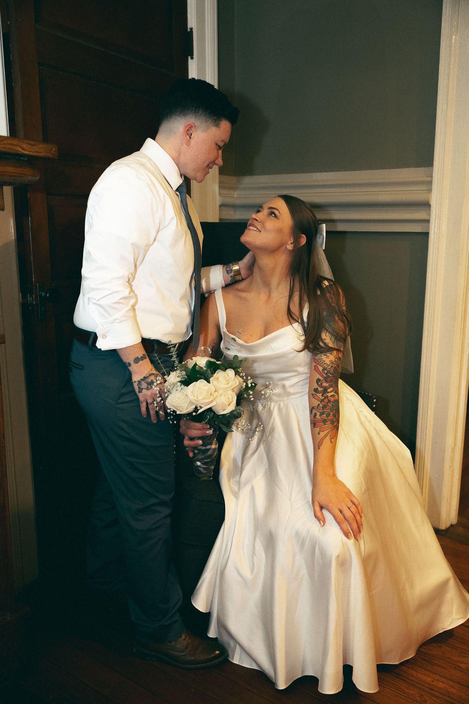 Newlywed couple sharing an intimate moment while holding white flowers in a dimly lit indoor setting.
