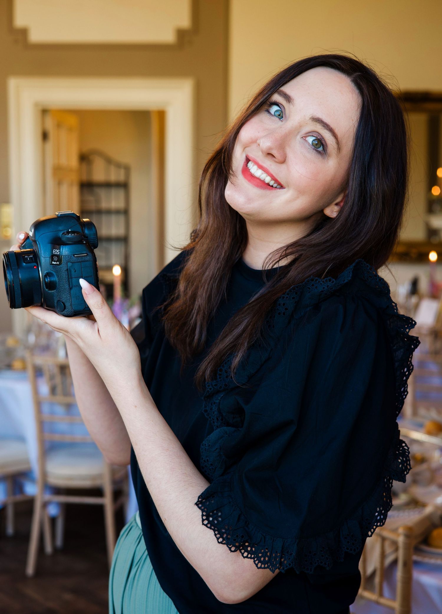 Smiling woman with dark hair holding a DSLR camera in an elegant event venue with table settings.