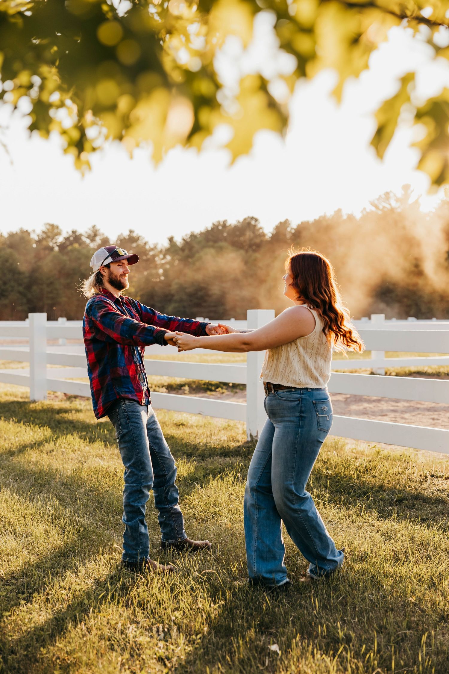 Two people in casual attire dance and hold hands in a sunlit field with white fence during golden hour.