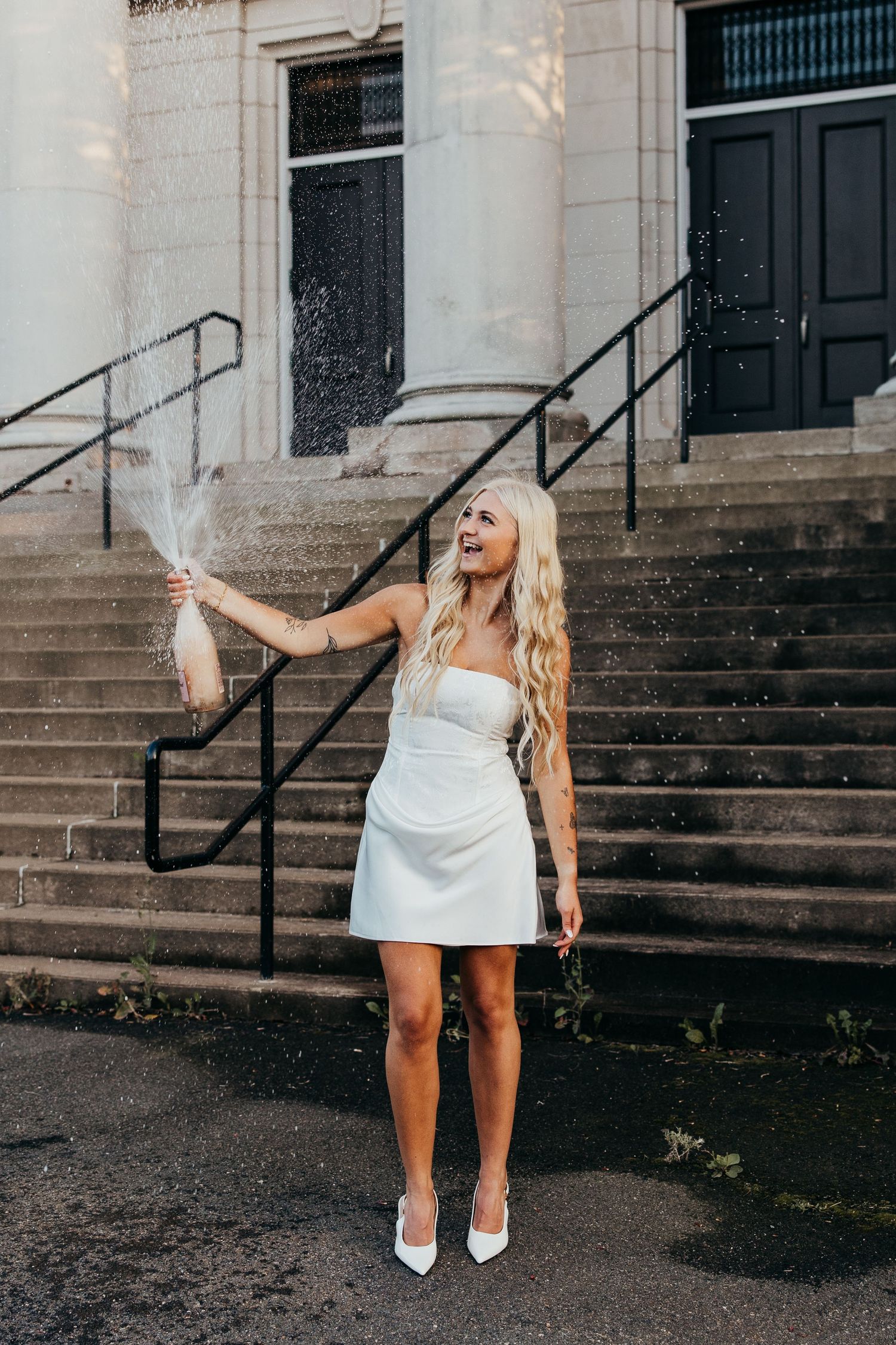 Smiling woman in a white mini dress and pointed white heels sprays champagne while celebrating on stone steps outside a columned building.