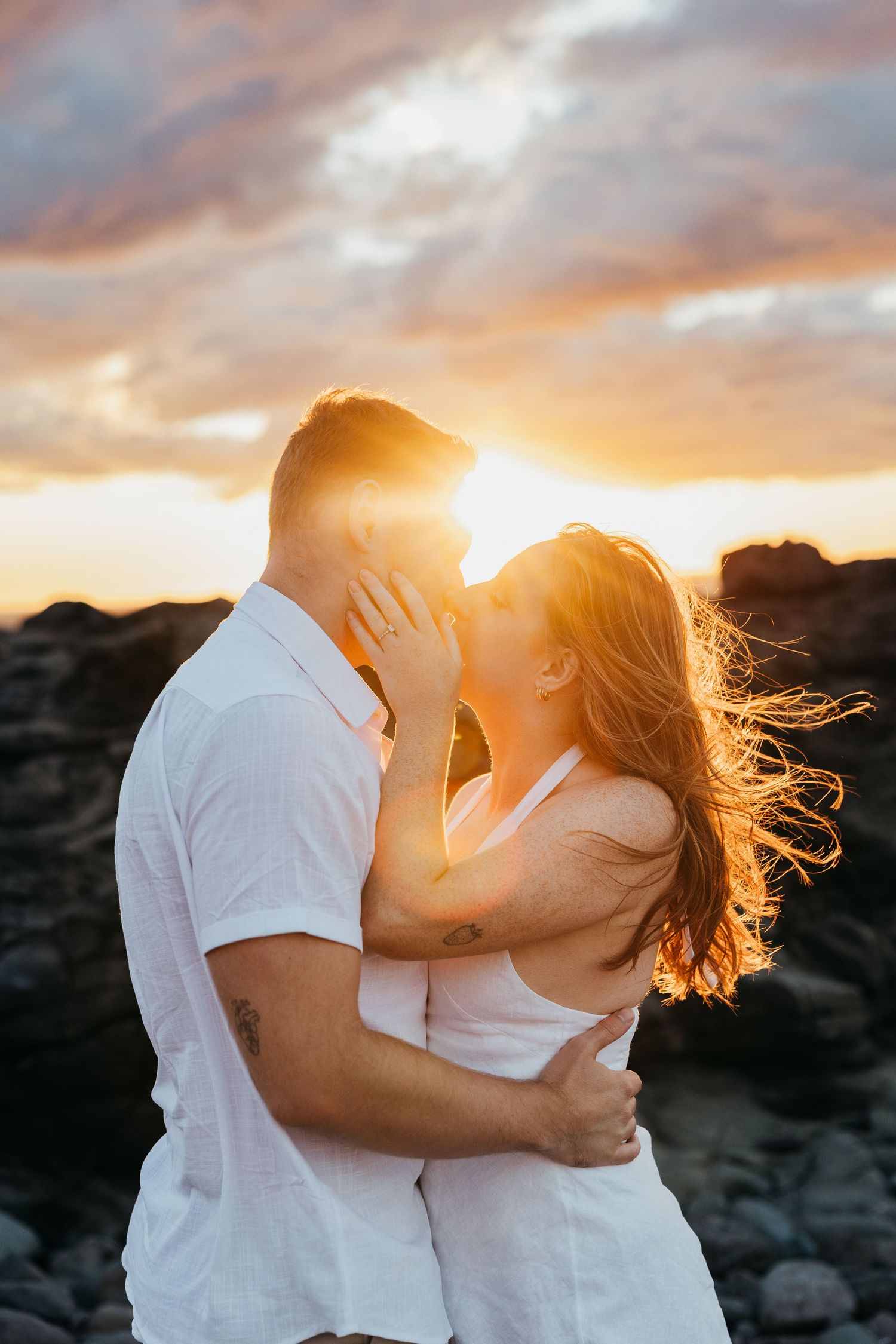 Couple kissing at sunset on rocky beach, golden light glowing behind them, woman's hair blowing in wind.