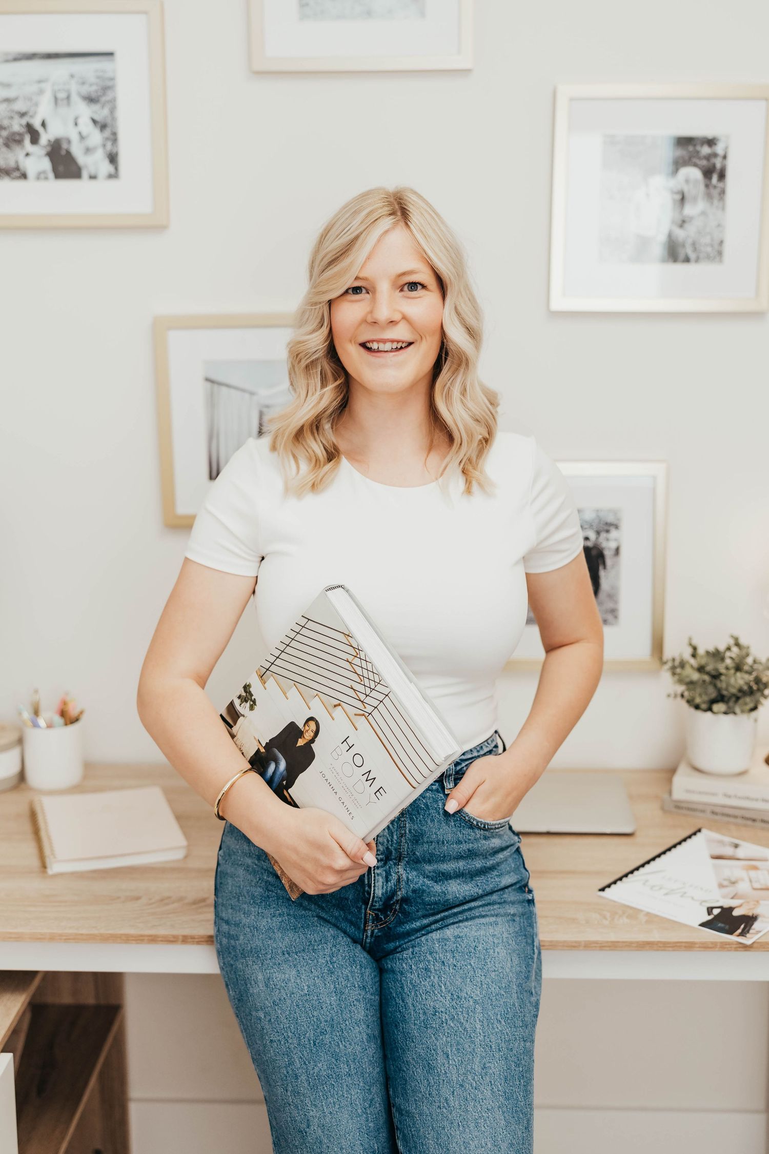 Woman in white t-shirt and jeans poses with books against wall with framed photos in bright interior setting.