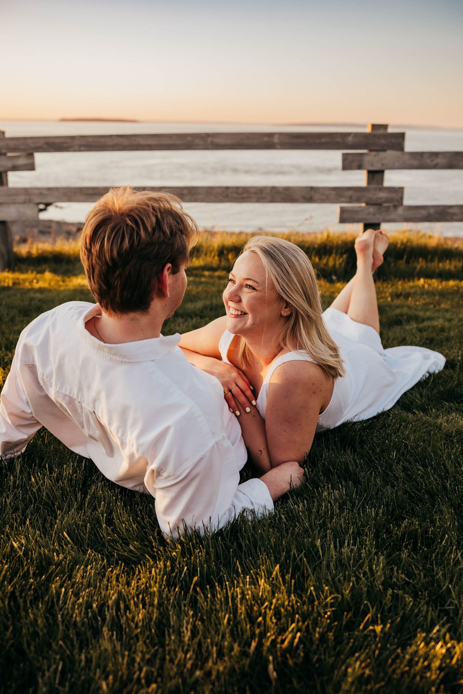 A couple lying in grass at sunset sharing a joyful moment near a wooden fence by the water.