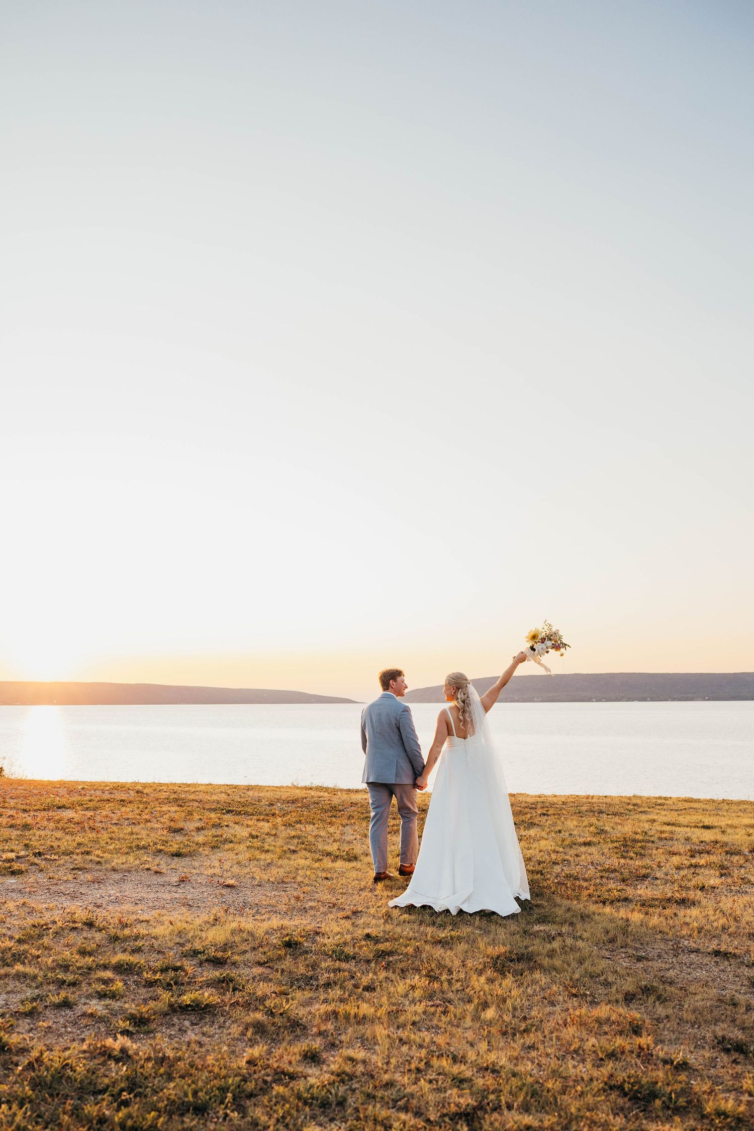 A couple in wedding attire walks across a golden hillside at sunset, creating a romantic silhouette against the sky.