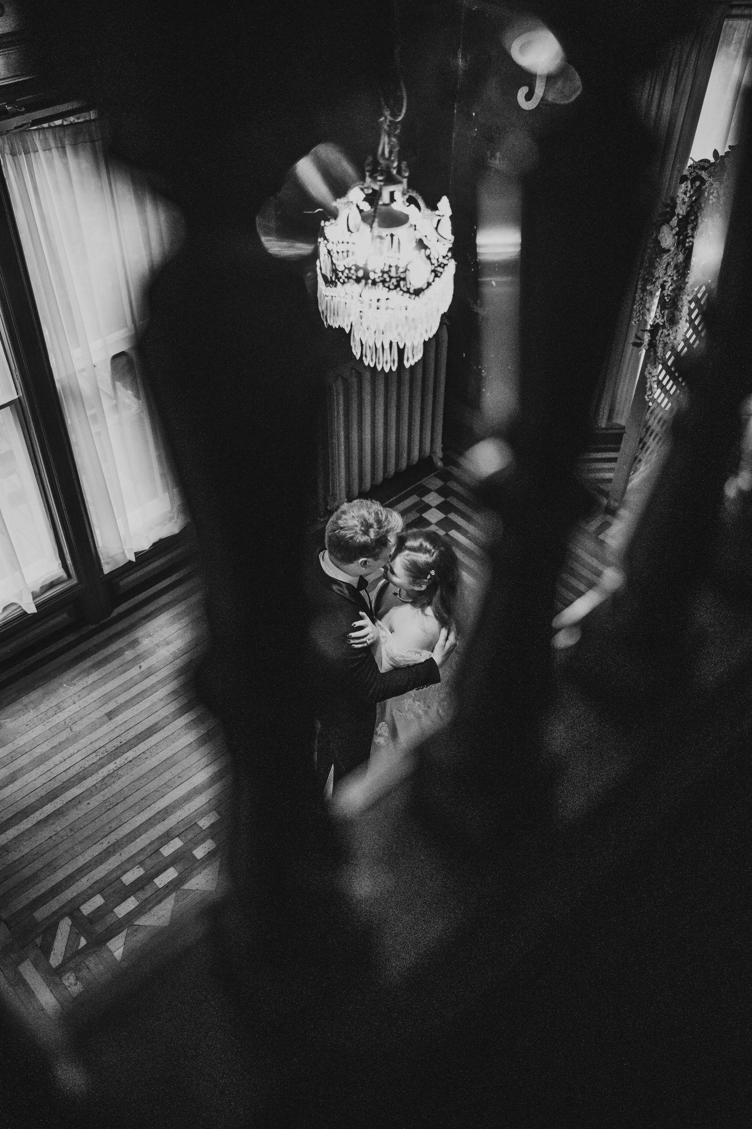 A black and white image shows a couple embracing beneath an ornate chandelier in a dimly lit room.