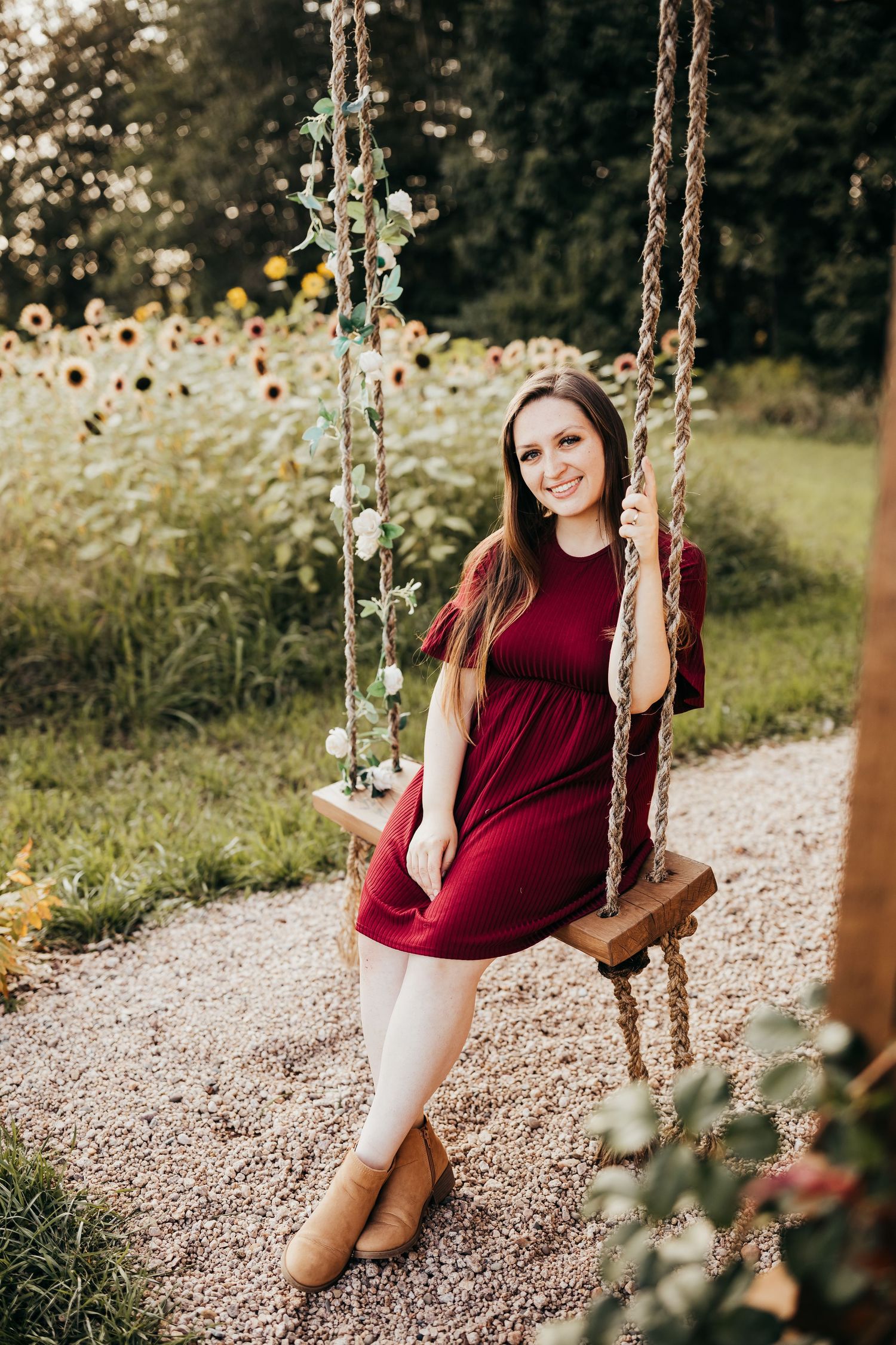 Portrait of someone in a burgundy dress sitting on a rustic rope swing with sunflowers in background.