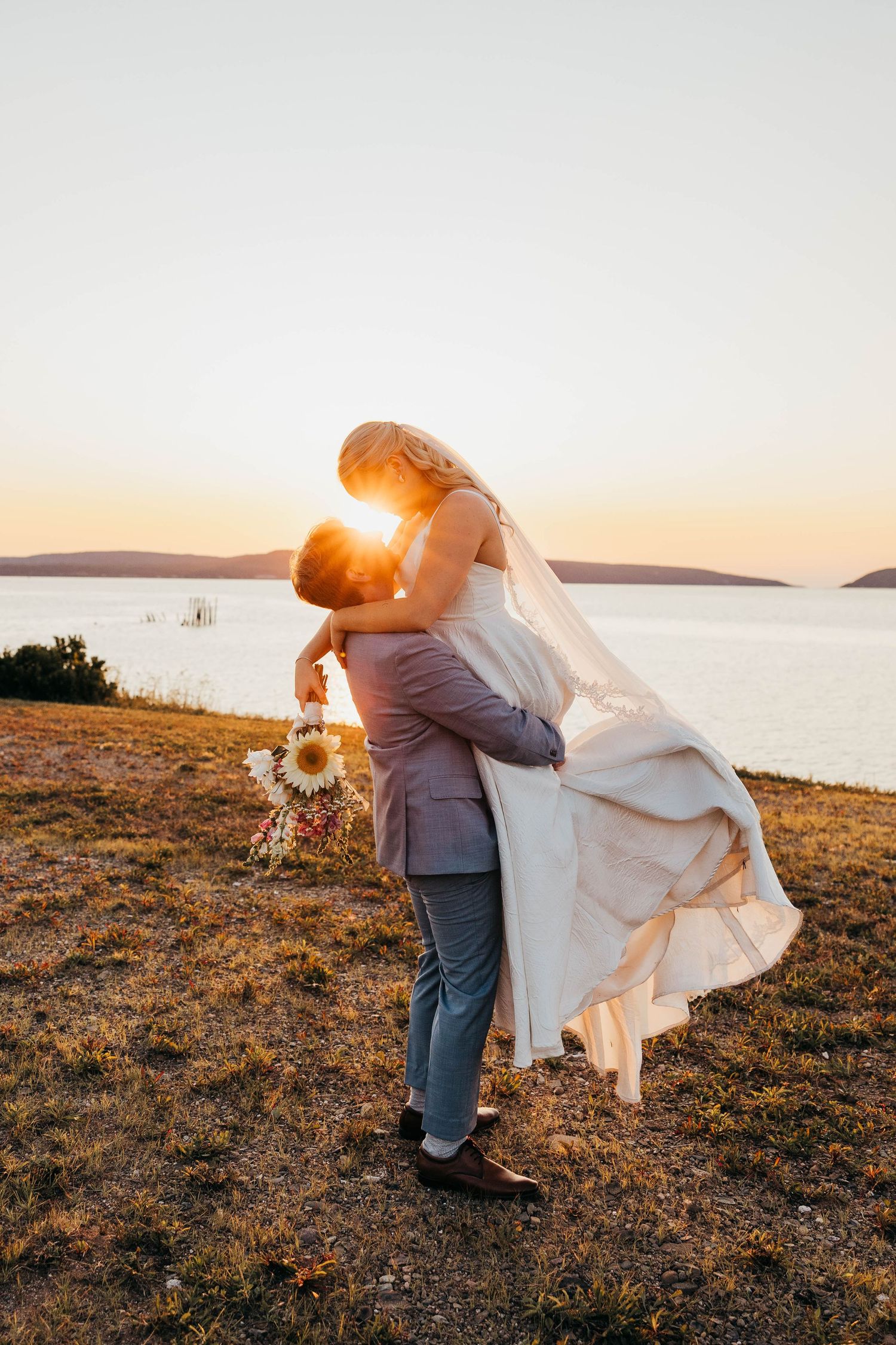 Newlyweds embrace on a hilltop during golden sunset near the ocean with the bride's dress flowing in the wind.