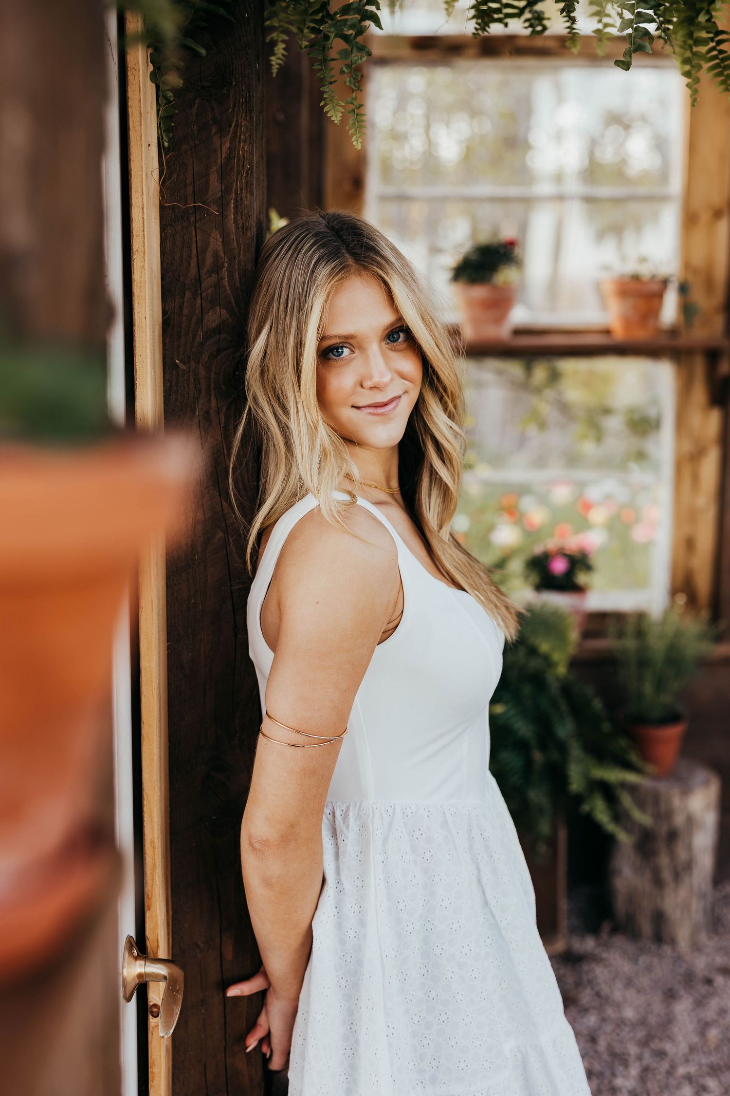 A woman in a white summer dress poses against a rustic door frame with potted plants in the background.