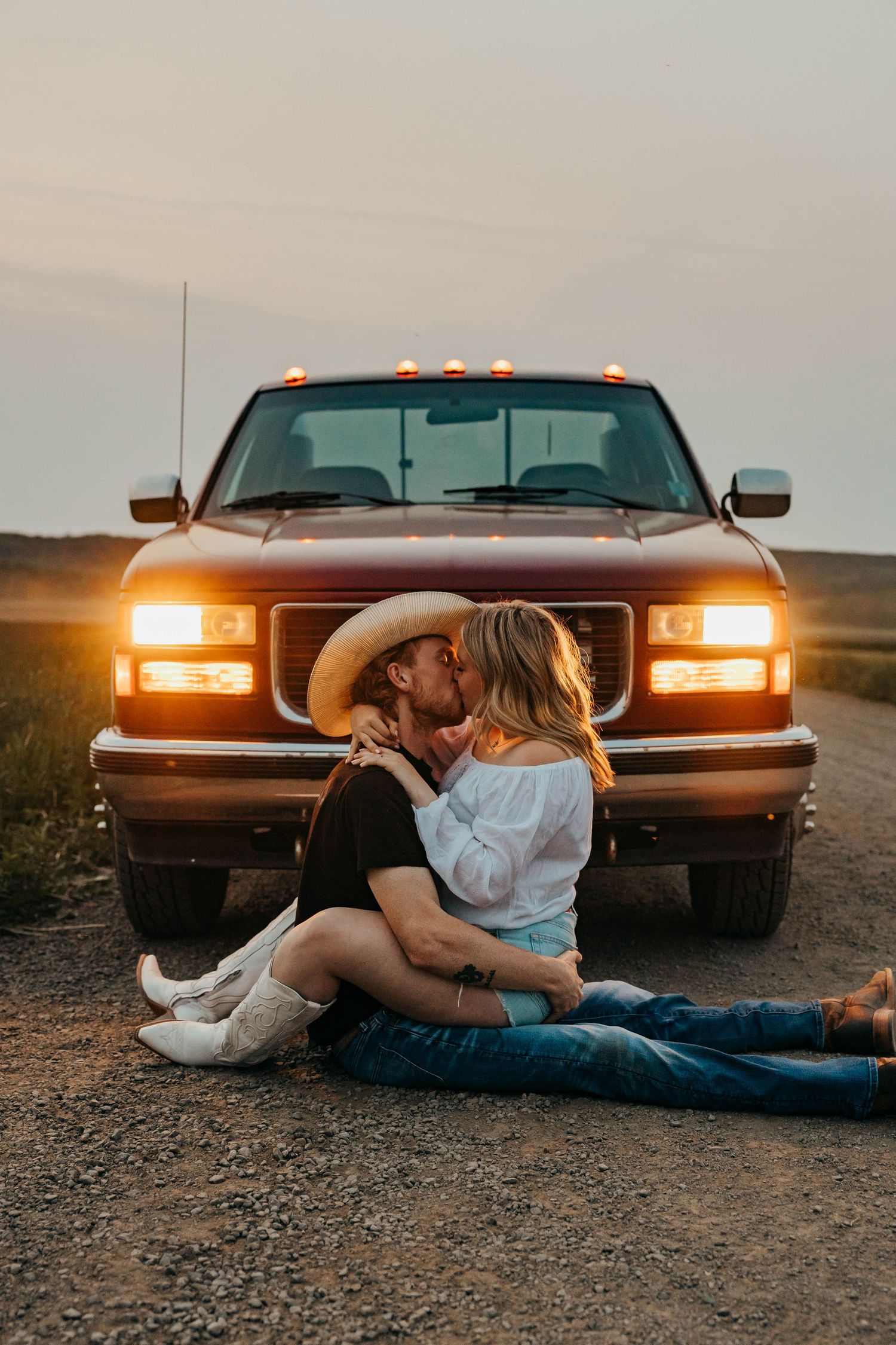 A romantic country scene with a couple embracing in front of a pickup truck at sunset with headlights glowing.