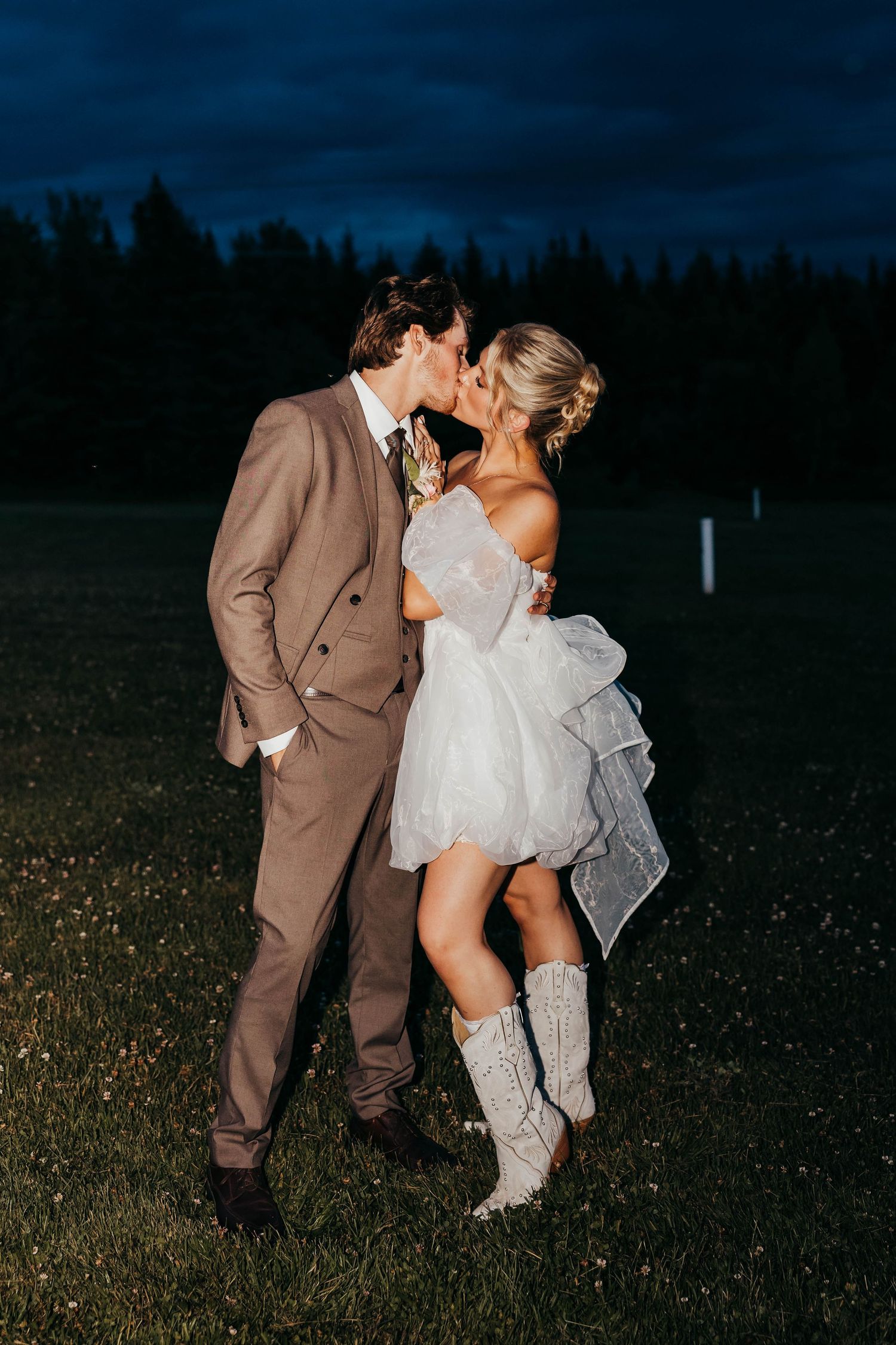 A romantic wedding couple embraces at dusk, the bride wearing cowboy boots and a short dress with the groom in a brown suit.